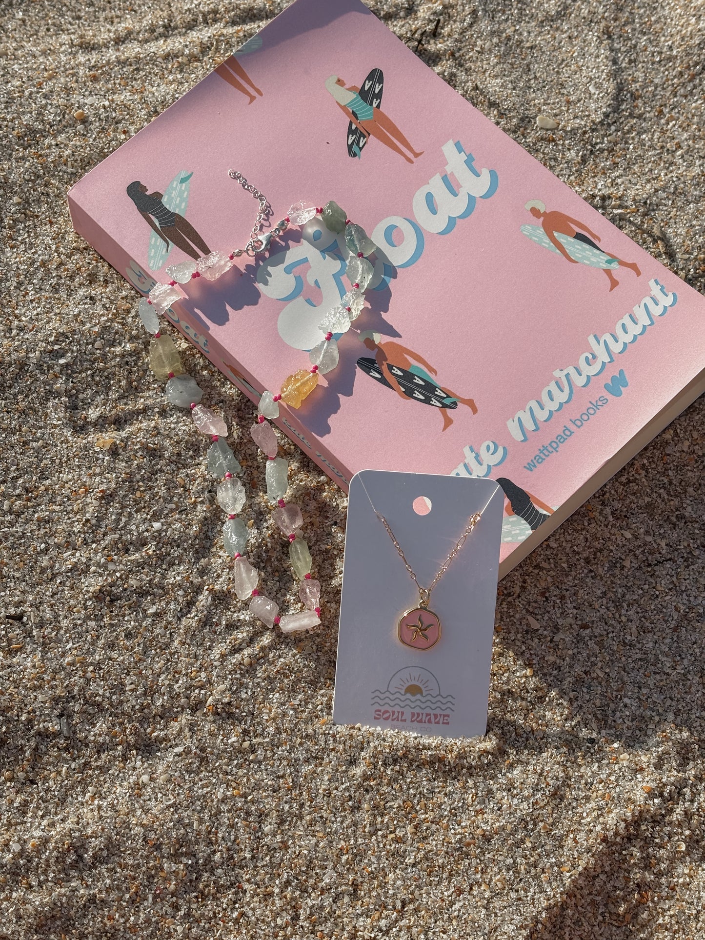 A flat-lay photo on the sand featuring a pink book titled “Float” with illustrated surfers on the cover. On top of the book is a colorful gemstone and simulated sea glass necklace with pink accent knots and a silver clasp. Next to the book is a gold-filled necklace with a round charm engraved with a starfish, displayed on a Soul Wave Beach Co. jewelry card.
