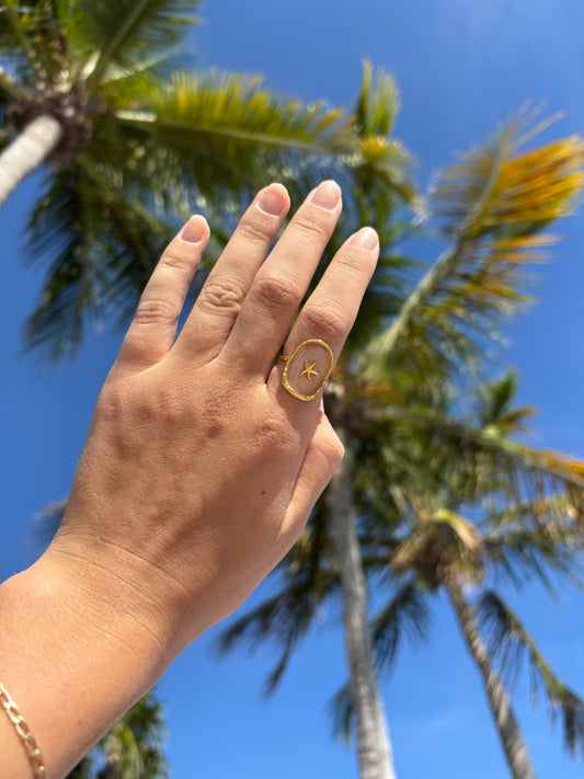 Close-up of a hand wearing the Coastal Keepsake Ring from Soul Wave Beach Co’s Ocean Heritage Collection. The ring features a 14k gold-plated band with a clear resin dome and a floating gold starfish charm inside. Photographed outdoors with a backdrop of blue sky and palm trees, this image highlights beach-inspired, handmade jewelry designed for ocean lovers. The ring embodies coastal elegance and tropical style, perfect for summer accessories, resort wear, and boho beach fashion. Ideal for customers search
