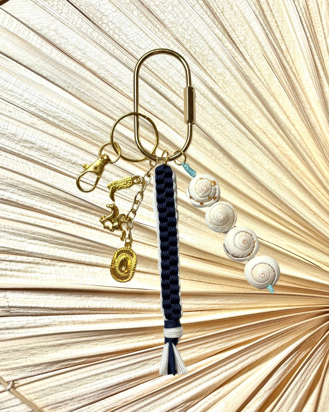 Flat lay of handmade Coastal Cowgirl keychain on a neutral palm fan background. Features spiral snail shell beads, navy and white knotted paracord cord, and gold western charms—cowboy boot, horse, and rope-etched medallion—on sturdy gold keyring hardware. Nautical cowgirl keychain for beach bags or purses. Western beachy charm gift for women, summer travel, rodeo events, or bachelorette party favors. Unique coastal boho accessory for keychains or bags.