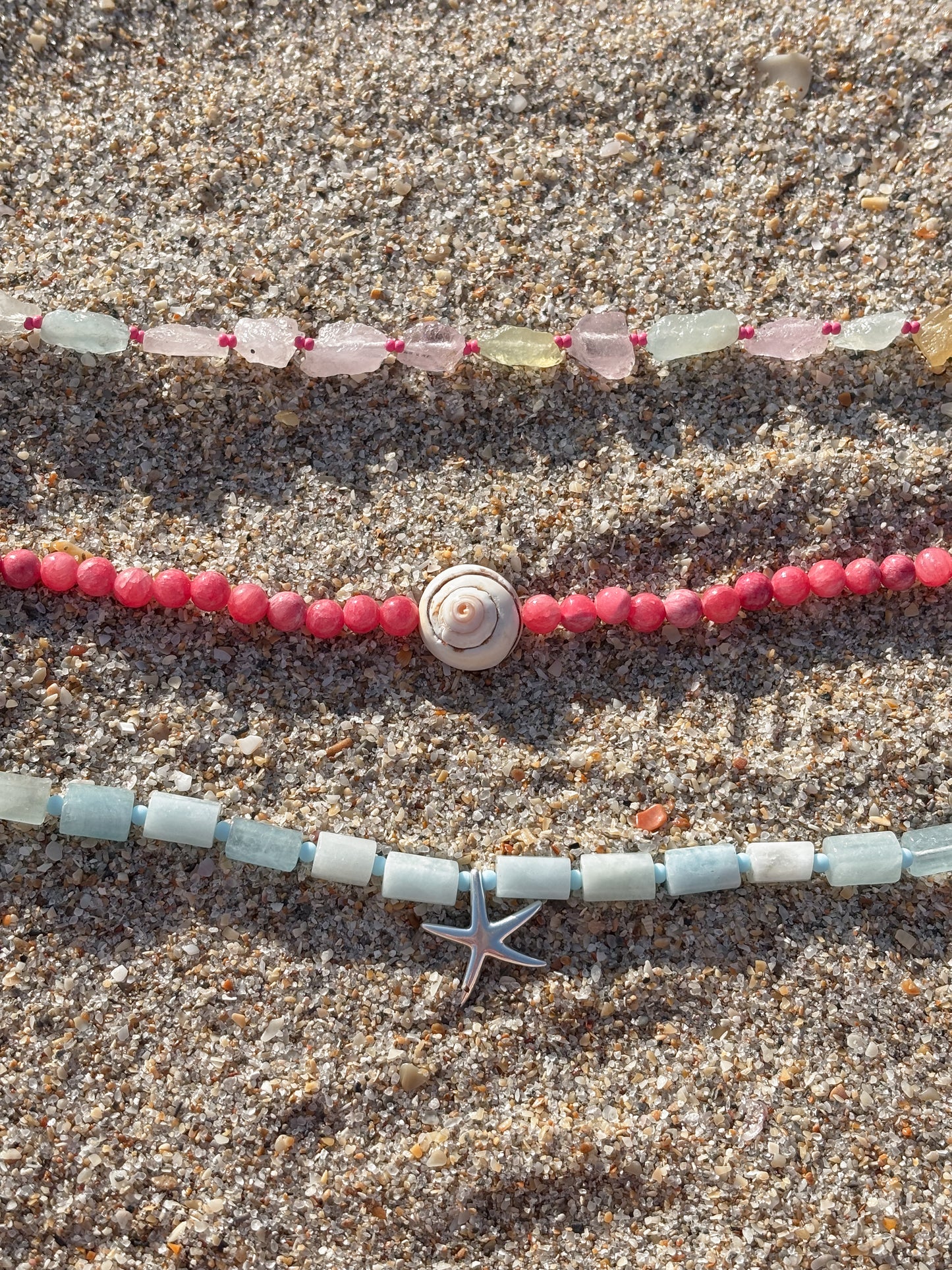 Three ocean-inspired beaded necklaces displayed on golden sand, perfect for layering. The top necklace features raw multicolored sea glass beads in soft pastels, hand-knotted on a vibrant pink cord. The middle necklace showcases smooth pink coral beads with a spiral shell centerpiece. The bottom necklace is made of square-cut aquamarine beads with a silver starfish charm, evoking a coastal, beachy aesthetic. A dreamy jewelry stack for summer, tropical vacations, and everyday ocean lovers.