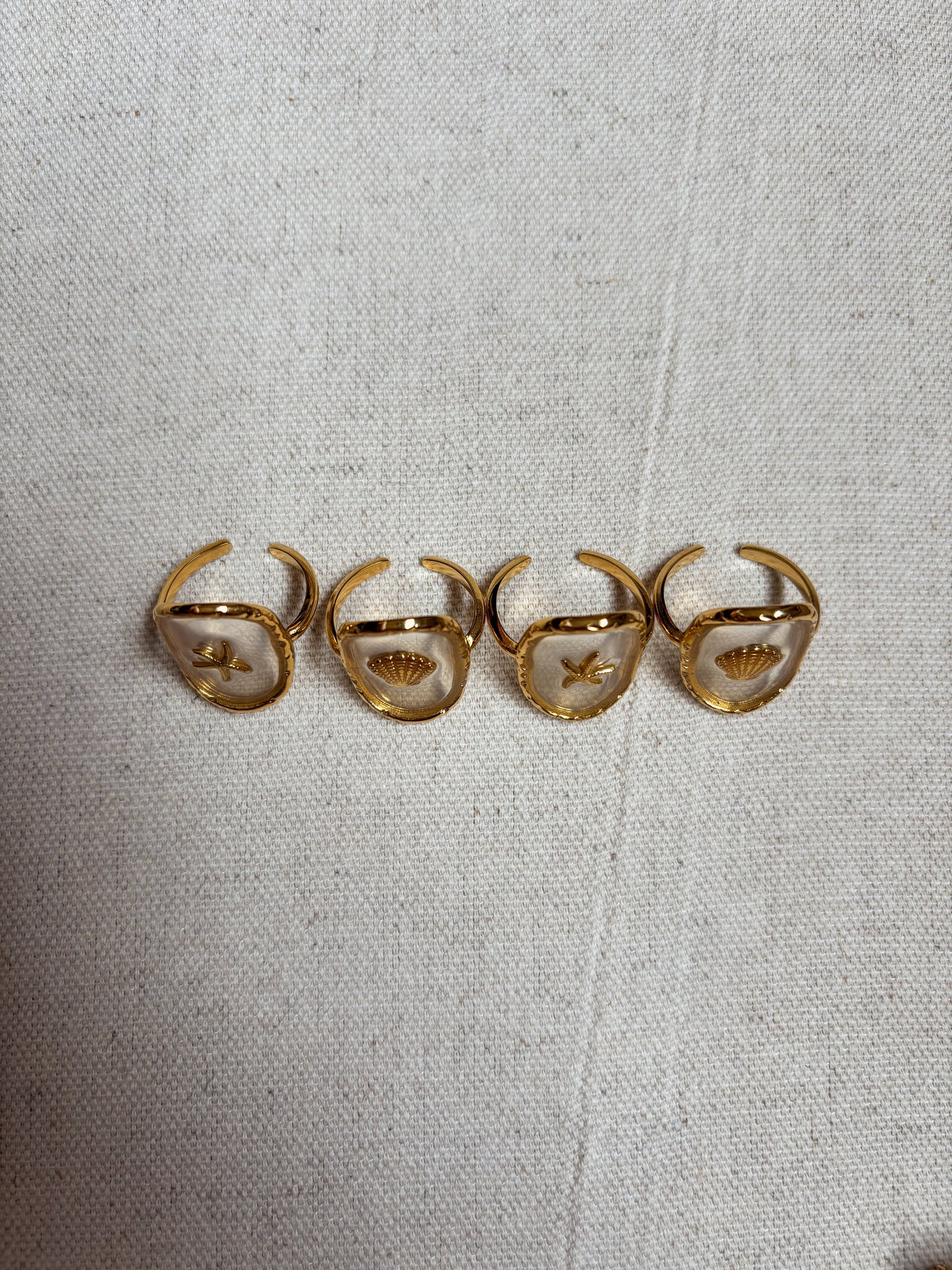 Top-down view of four gold-plated coastal rings from Soul Wave Beach Co’s Ocean Heritage Collection, displayed on a natural linen surface. Each adjustable open-band ring features a clear resin bezel with a miniature starfish or seashell charm inside, capturing a beachy, ocean-inspired aesthetic. These handmade beach rings reflect boho minimalism and coastal elegance, perfect for summer outfits, seaside jewelry lovers, and tropical vacation accessories. Ideal for customers searching for adjustable gold beach