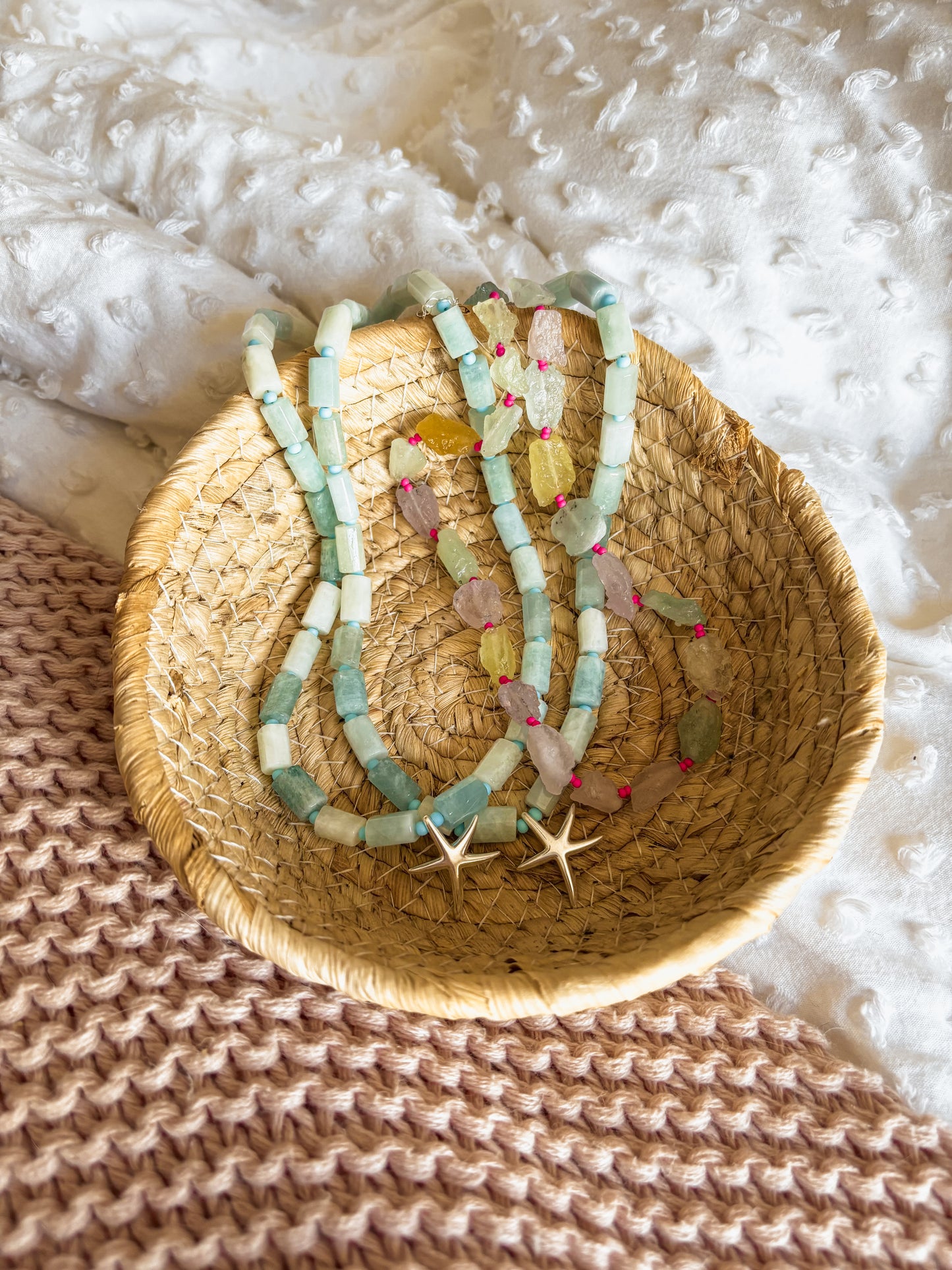 A woven seagrass basket holds two handmade beachy necklaces with natural Amazonite beads, raw gemstones, and pink knot accents. Gold starfish charms add a coastal touch. Styled on a textured knit blanket with soft white bedding, this boho, ocean-inspired jewelry radiates tropical vibes.