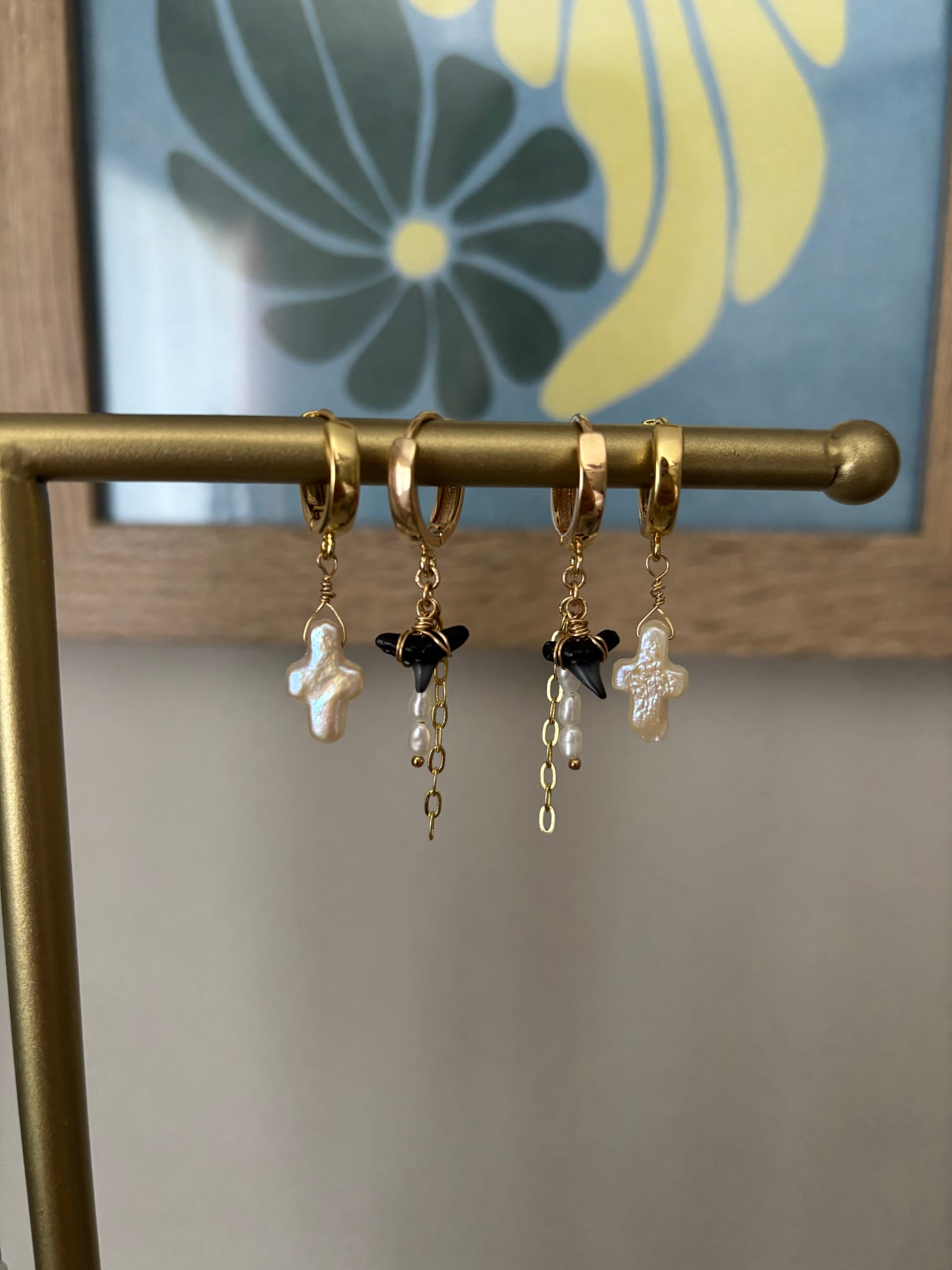 Two pairs of mismatched gold huggie hoop earrings displayed on a brass jewelry bar. Features two pearl cross earrings and two black shark tooth earrings with freshwater pearls and gold chain dangles. All earrings are handcrafted with 14k gold-filled wire and 18k gold-plated hoops. Designed by Soul Wave Beach Co., this coastal jewelry blends beachy elegance with bold ocean vibes. Set against a floral art background for a modern tropical aesthetic.
