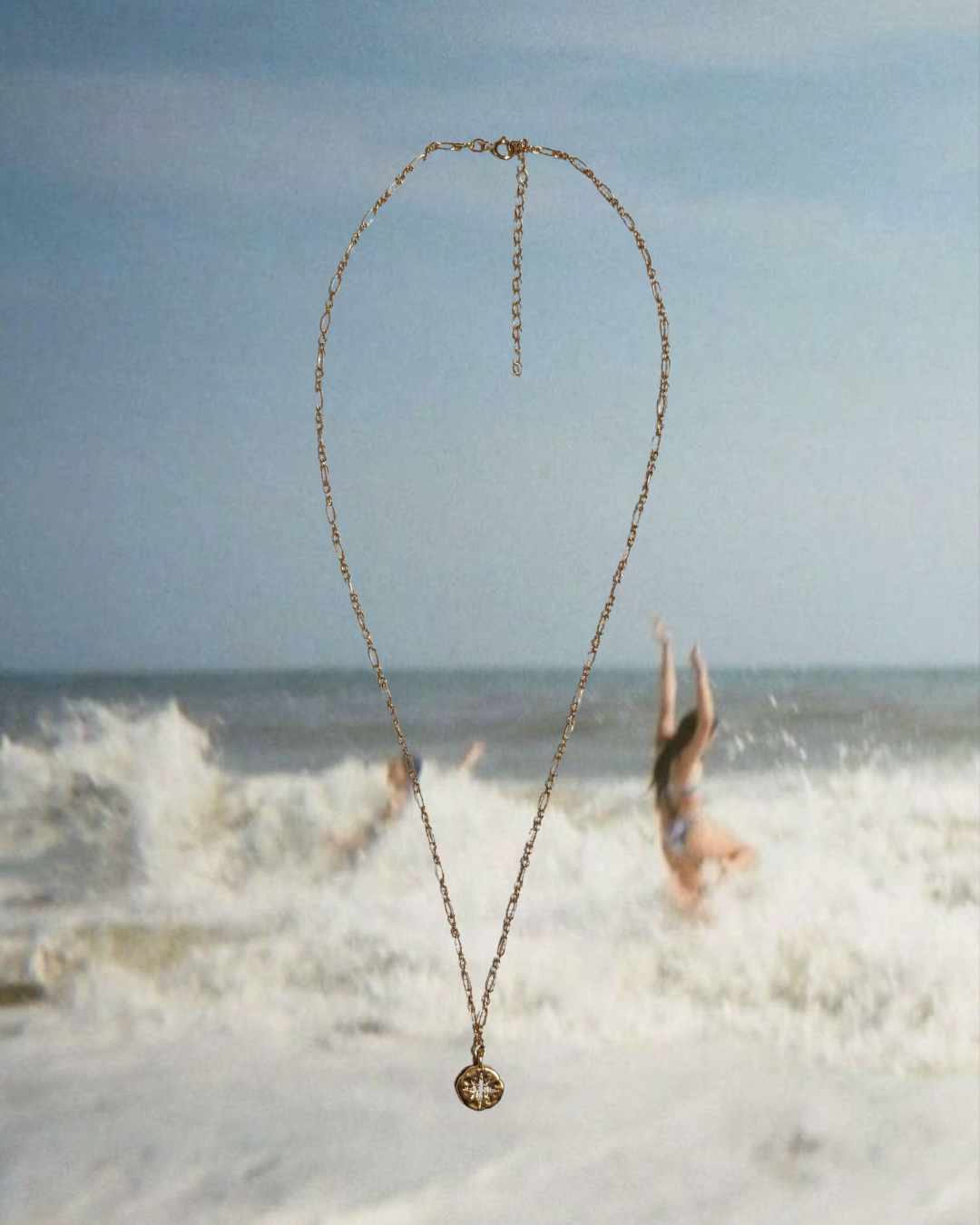 A delicate gold chain necklace featuring a small, textured shell charm with a sparkling crystal center is displayed against a nostalgic, sun-soaked beach backdrop. The background shows two women mid-laughter, splashing in ocean waves—capturing carefree summer joy. This piece from Soul Wave Beach Co’s Ocean Heritage collection channels vintage coastal energy, golden hour glow, and effortless beach style, perfect for layering or wearing solo for a timeless seaside look.