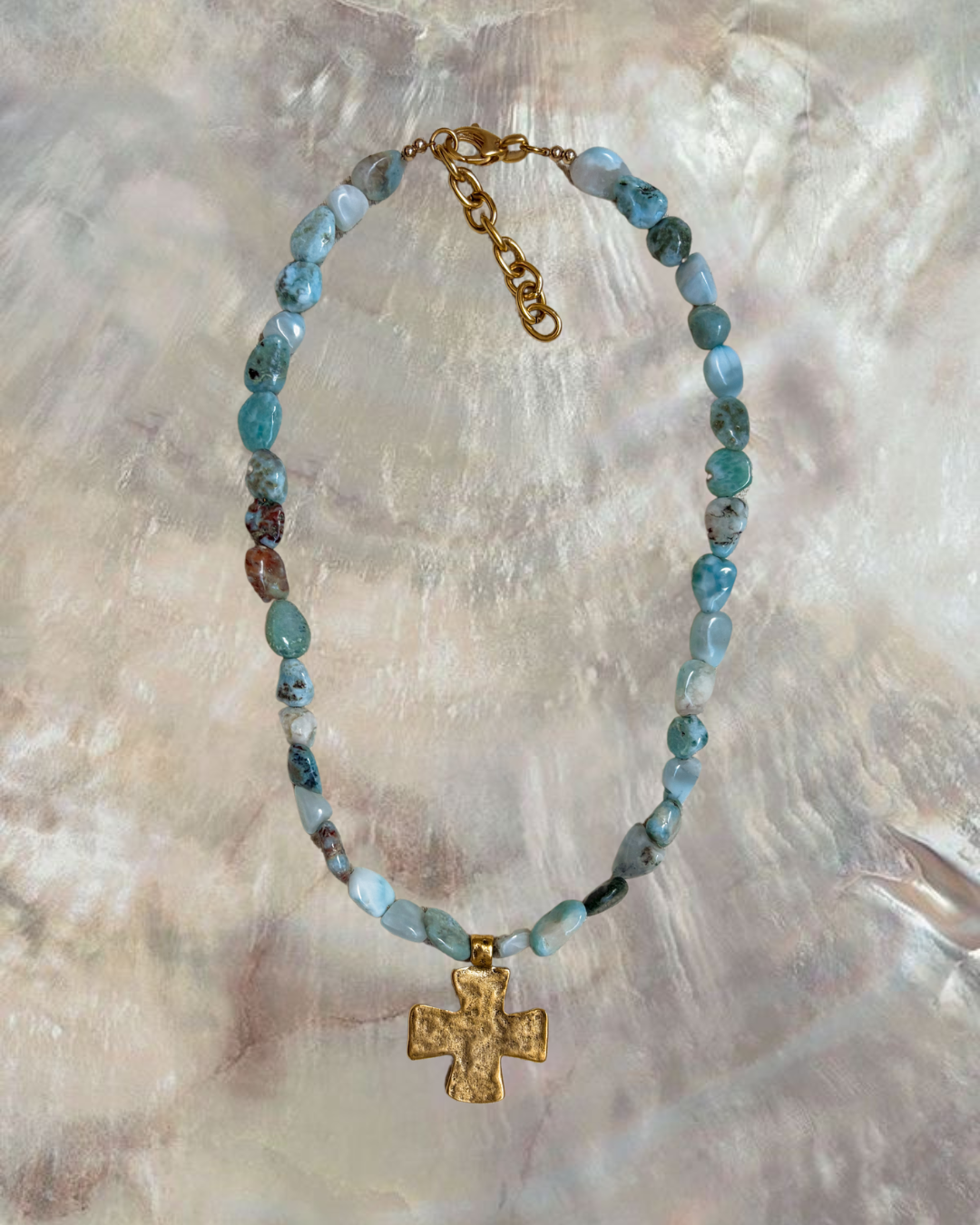A chunky larimar beaded necklace with a textured gold cross pendant and adjustable gold extender chain lies against an iridescent shell background. The natural blue larimar stones vary in tone, evoking ocean waves and coastal rock textures. Designed by Soul Wave Beach Co, this piece from the Ocean Heritage collection channels tropical serenity and coastal spirituality, blending beach-inspired elegance with earthy, handcrafted charm.