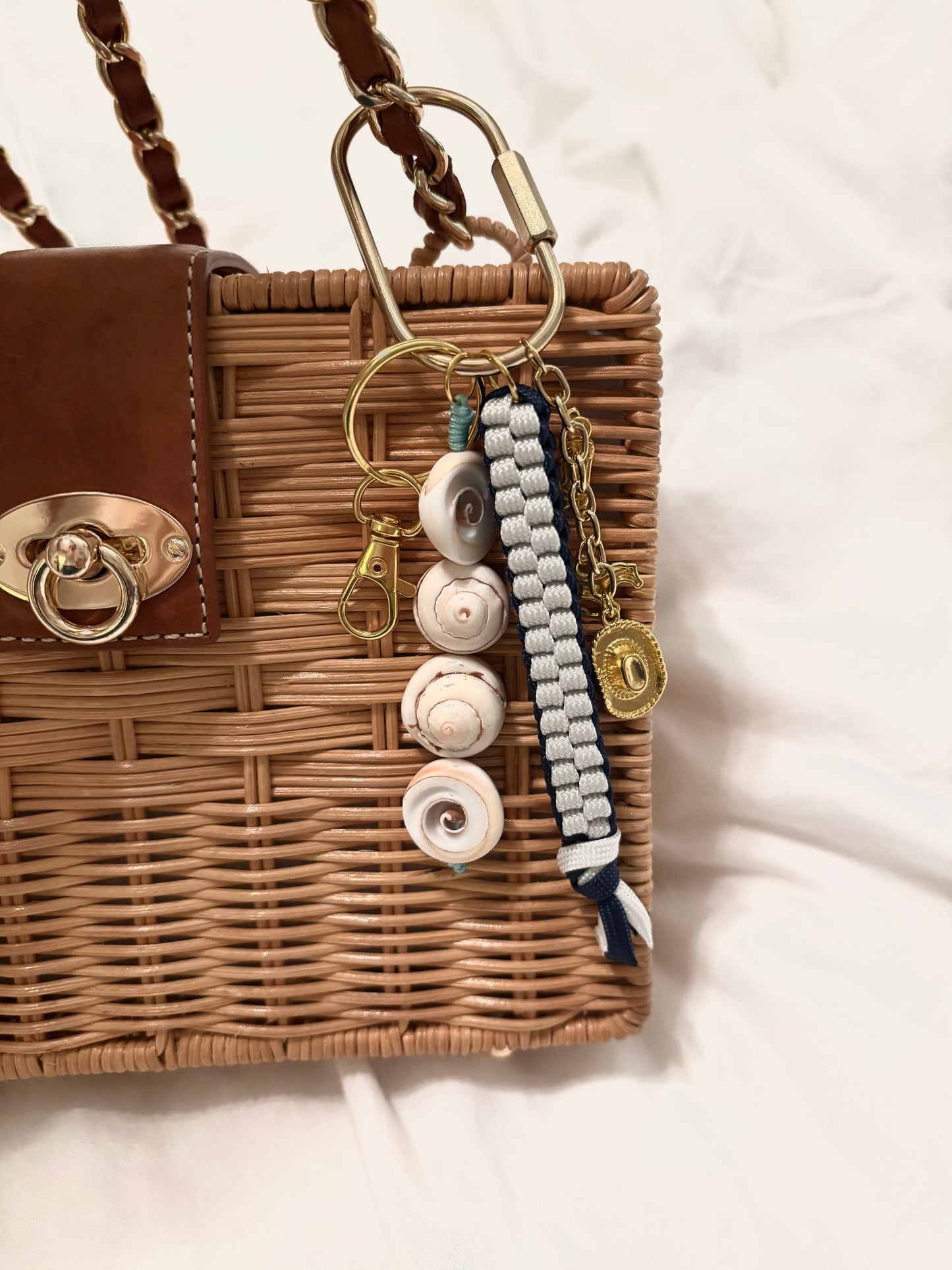Coastal Cowgirl bag charm hanging on a rattan woven purse, featuring real spiral seashell beads, navy and white knotted paracord cord, and gold western charms including a cowboy boot, galloping horse, and engraved medallion. Beachy cowgirl keychain and boho western bag charm, perfect for coastal cowgirl gifts, beach vacations, rodeo season, or summer accessory styling. Handmade beach charm adds ocean-inspired flair to any tote or handbag.