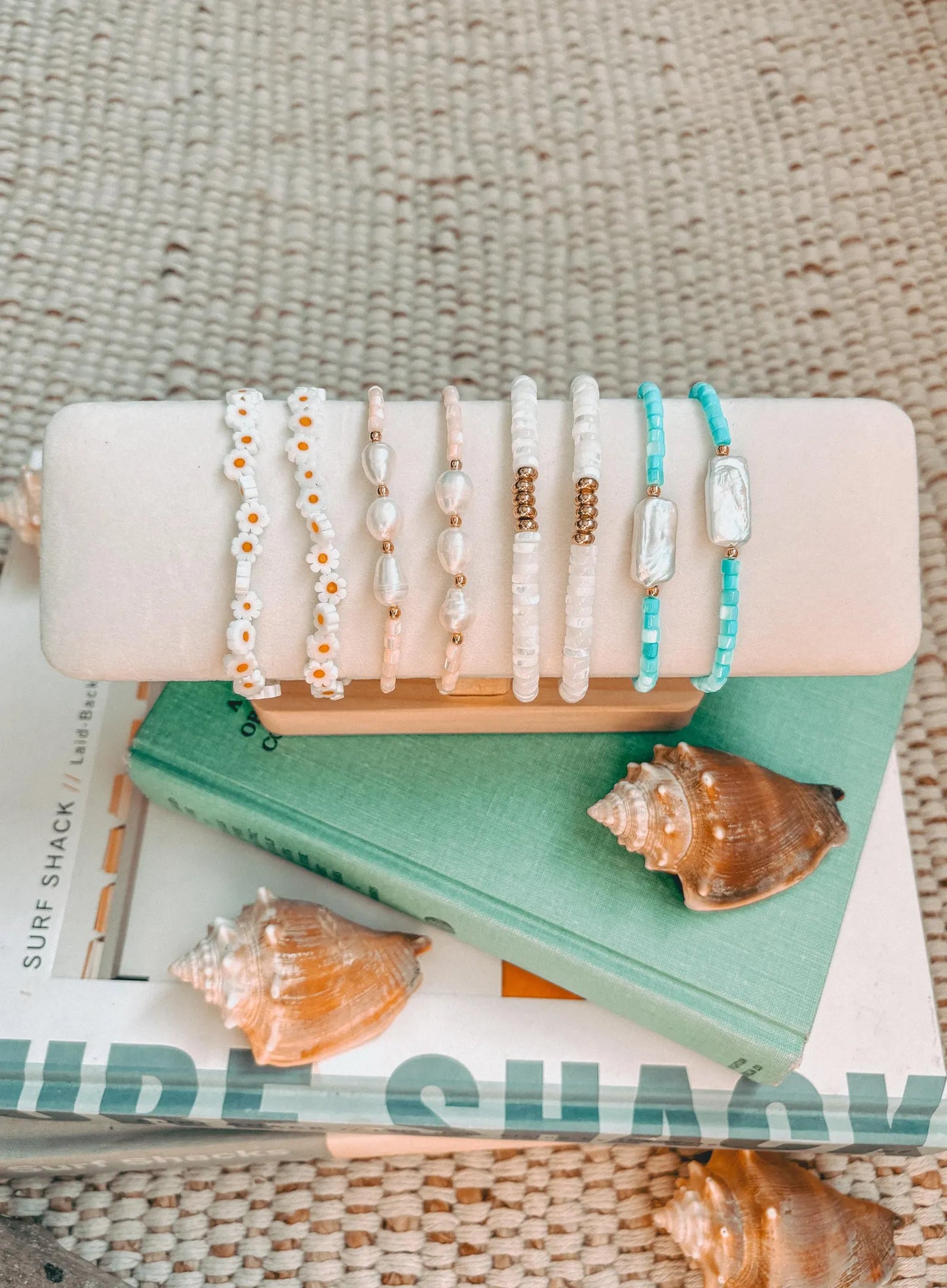 Display of beach-inspired jewelry featuring delicate bracelets with daisy designs, freshwater pearls, white and turquoise beads, and gold accents. Set against a coastal aesthetic with seashells, textured fabric, and beach-themed books, the image captures the essence of a sunny, seaside vibe. Perfect for showcasing handmade coastal jewelry collections