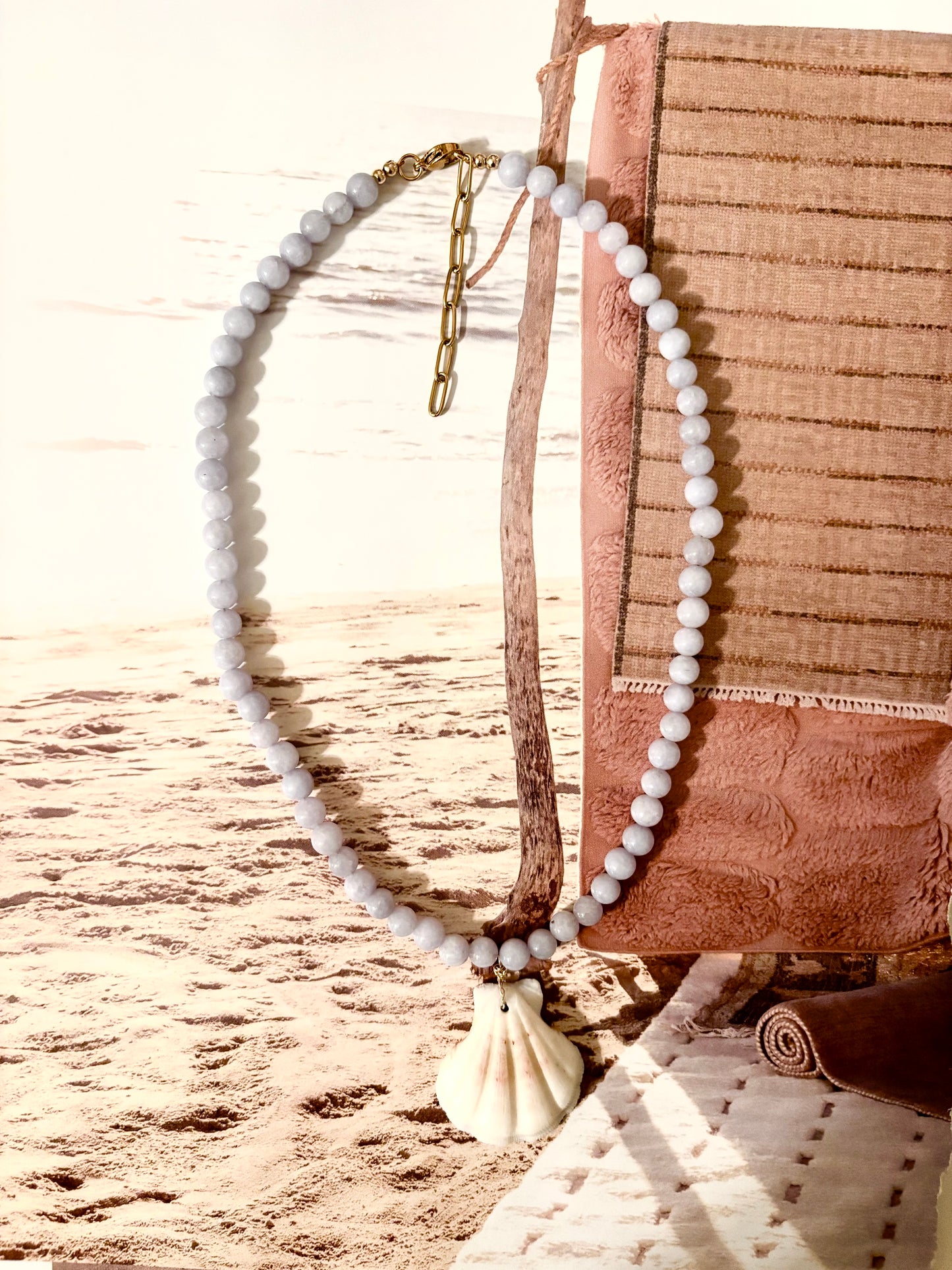 Light blue beaded necklace with natural scallop shell pendant, photographed against a beachy backdrop with sand and driftwood. Handmade by Soul Wave Beach Co, this ocean-inspired piece features blue aquamarine beads, a 18k gold-filled clasp, and adjustable extender chain. A soft coastal necklace perfect for layering or making a relaxed summer statement.