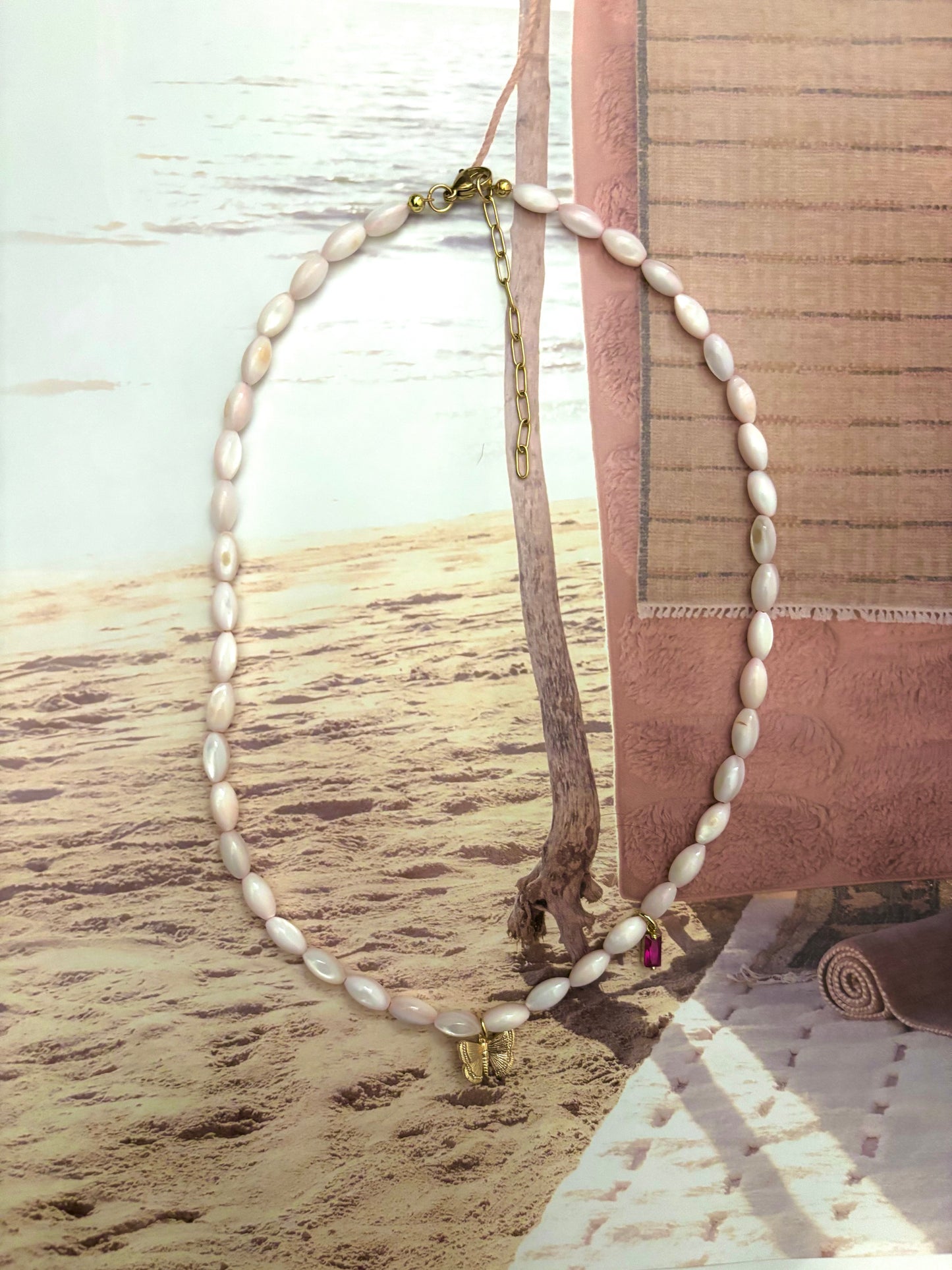 Blush pink coral bead necklace with gold butterfly charm and hot pink crystal accent, styled on a sandy beach backdrop with ocean in the distance. Features a 18k gold-plated clasp and extender chain. Handmade by Soul Wave Beach Co, this soft and coastal necklace offers a feminine, beachy touch—perfect for summer layering or dreamy resort styling.