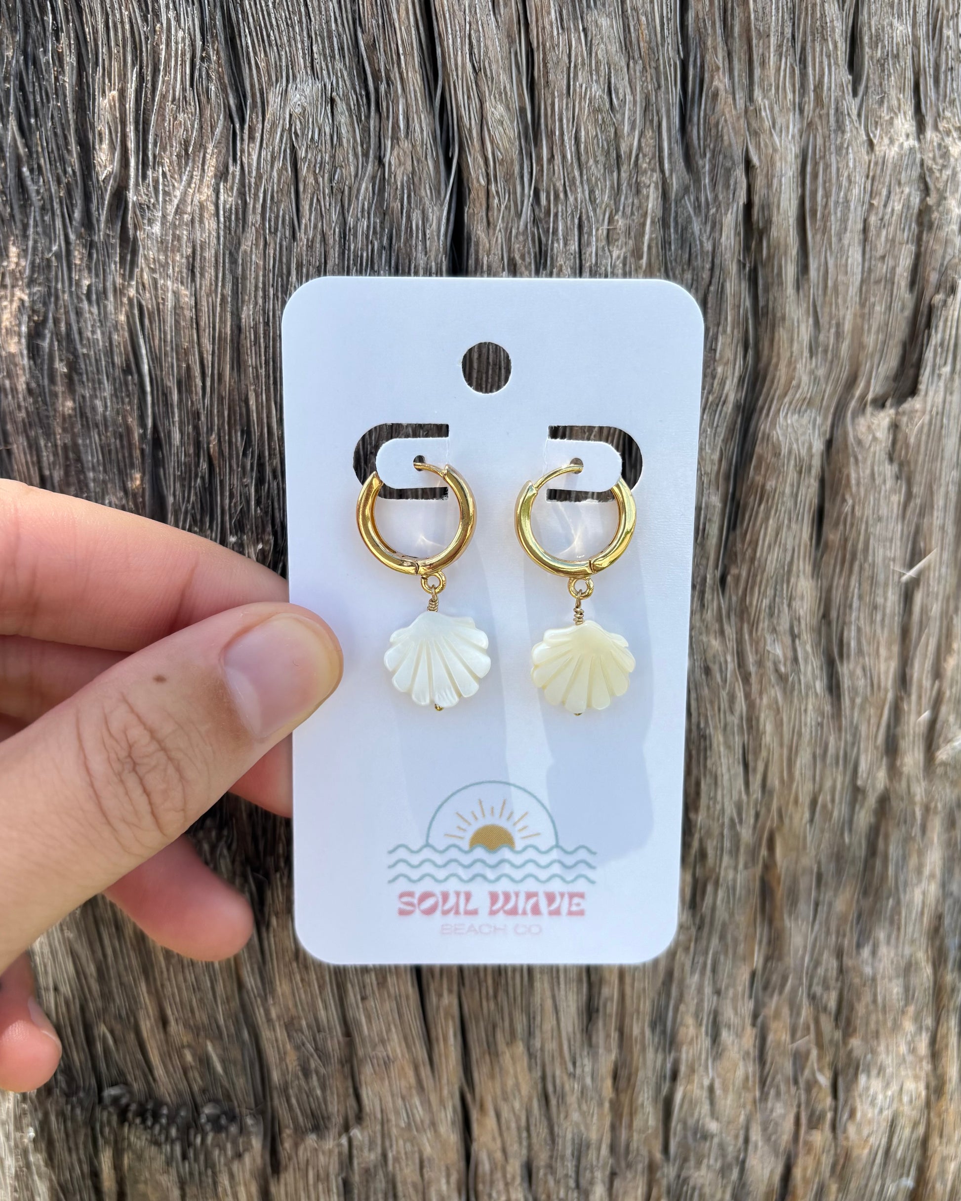 Hand holding a pair of mother of pearl shell earrings on a Soul Wave Beach Co. earring card. Earrings feature white carved shell charms and 18k gold-plated huggie hoops with 14k gold-filled wire. Photographed against weathered driftwood for a rustic coastal look. Lightweight beach earrings perfect for tropical vacations, handmade ocean jewelry with gold and pearl tones.