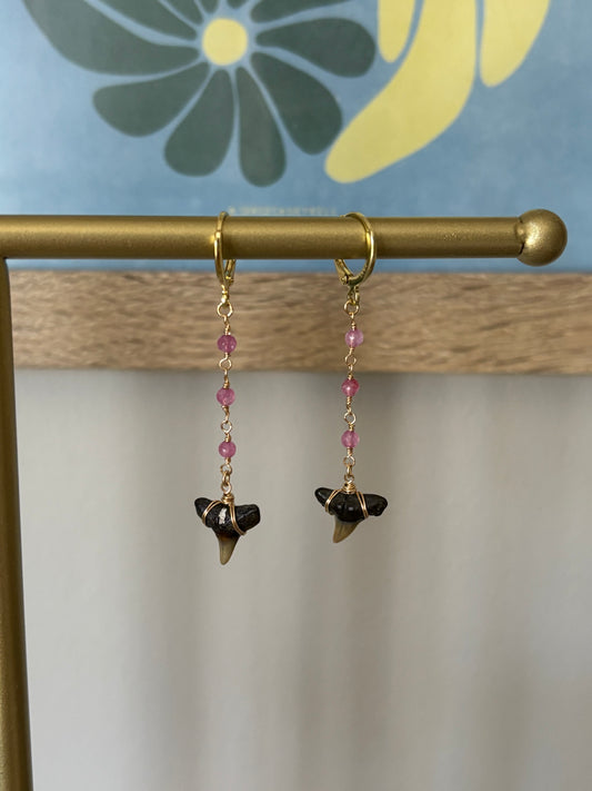 Close-up of pink glass beaded shark tooth earrings with fossil teeth wire-wrapped in 14k gold-filled wire and hanging from gold-filled hoops. Handmade by Soul Wave Beach Co., these earrings blend beachy charm and bold coastal energy. Lightweight and ideal for summer outfits, tropical vacations, or surf culture fashion. Displayed on a gold bar with floral background for tropical vibes.