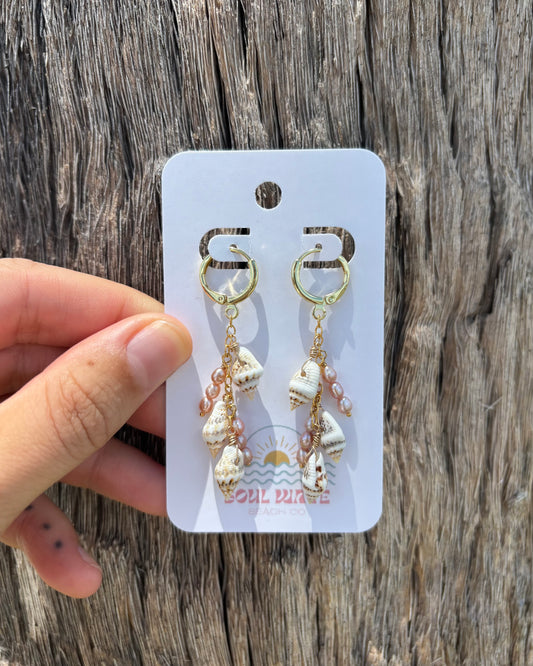 Beachy spiral shell and freshwater pearl dangle earrings on branded Soul Wave Beach Co. card. Handmade with natural spiral seashells and purplish-pink pearls, wire-wrapped in 14k gold-filled sterling silver. Coastal boho ocean-inspired jewelry, lightweight and beach chic. Perfect for summer, beach weddings, tropical vacations, or everyday resort wear. Photographed on rustic driftwood for natural seaside vibe.