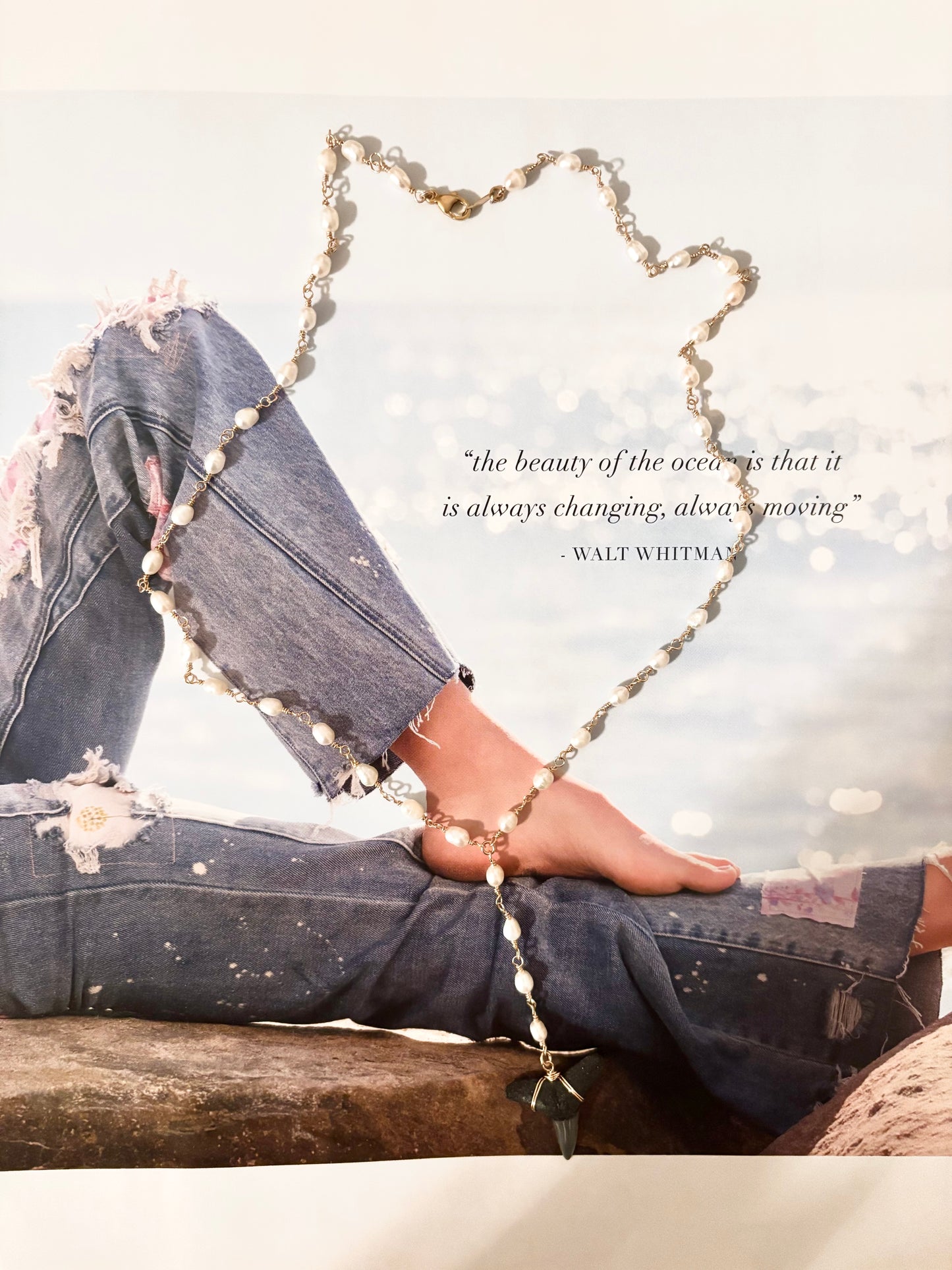 Lariat necklace with hand-wrapped freshwater pearls and a fossilized shark tooth pendant, styled on top of a beachy lifestyle magazine page featuring a model in ripped denim by the ocean. The necklace is made with 14k gold filled sterling silver, 18 inches with a 3-inch drop. Perfect for coastal jewelry lovers, beachy accessories, and ocean-inspired fashion.