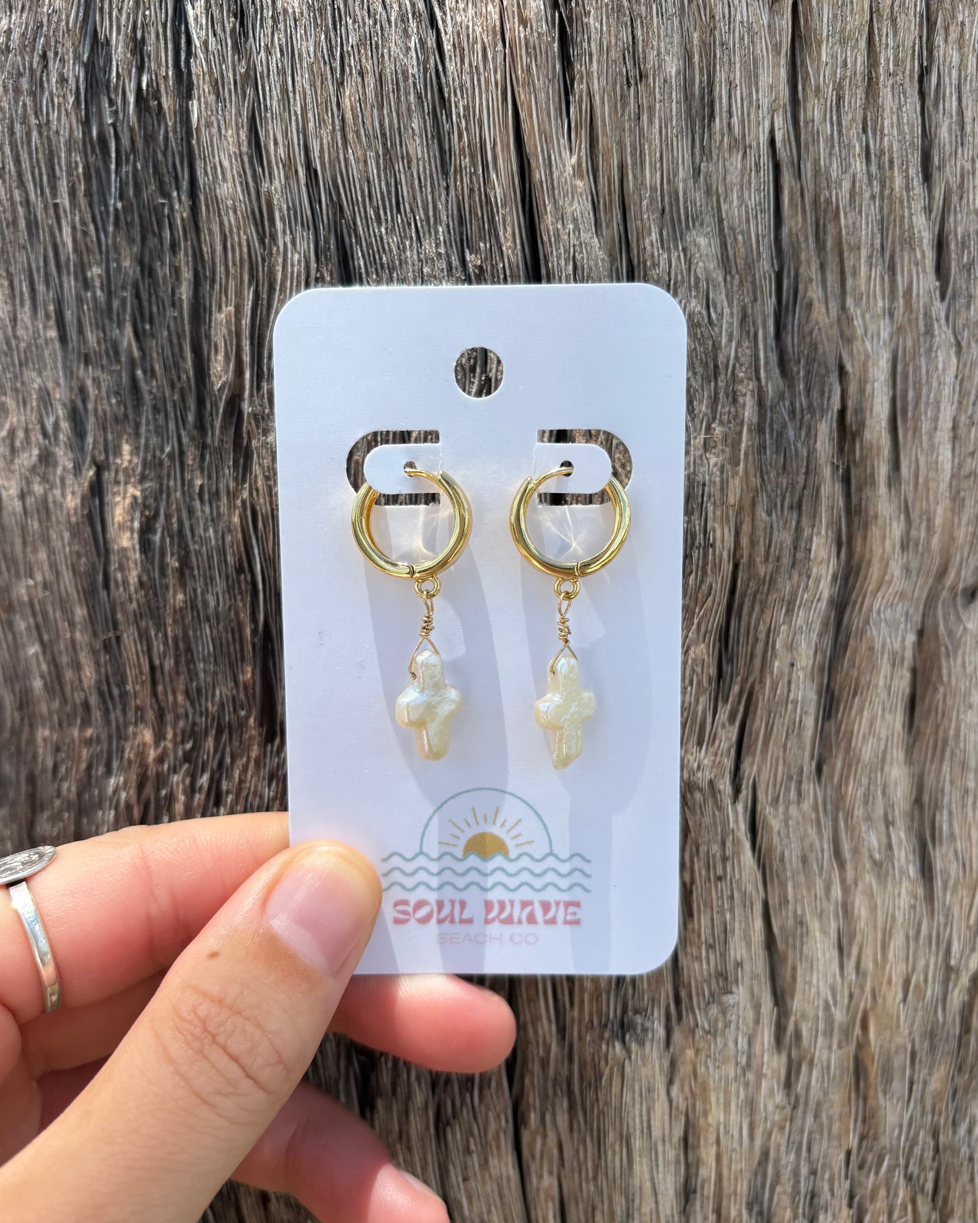 Close-up of pearl cross earrings held by hand on Soul Wave Beach Co. earring card. Features cross-shaped pearls suspended from 18k gold-plated huggie hoops and detailed with 14k gold-filled wire. Photographed on rustic driftwood for a natural seaside vibe. Handmade gold and pearl earrings perfect for beach brides, gifting, or daily wear.