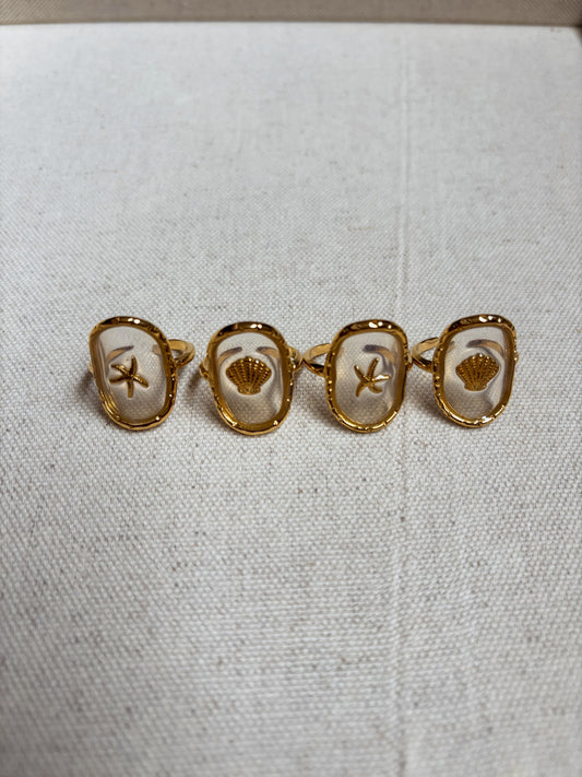A set of four gold-plated coastal-themed rings from Soul Wave Beach Co’s Ocean Heritage Collection displayed on a neutral linen background. Each ring features a unique beach-inspired charm—two starfish and two seashells—encased in a clear resin oval bezel. These handmade, beachy statement rings blend ocean-inspired design with boho minimalism, perfect for summer jewelry lovers, beachwear accessorizing, and coastal fashion. Ideal for customers seeking ocean rings, starfish and shell jewelry, and handcrafted 