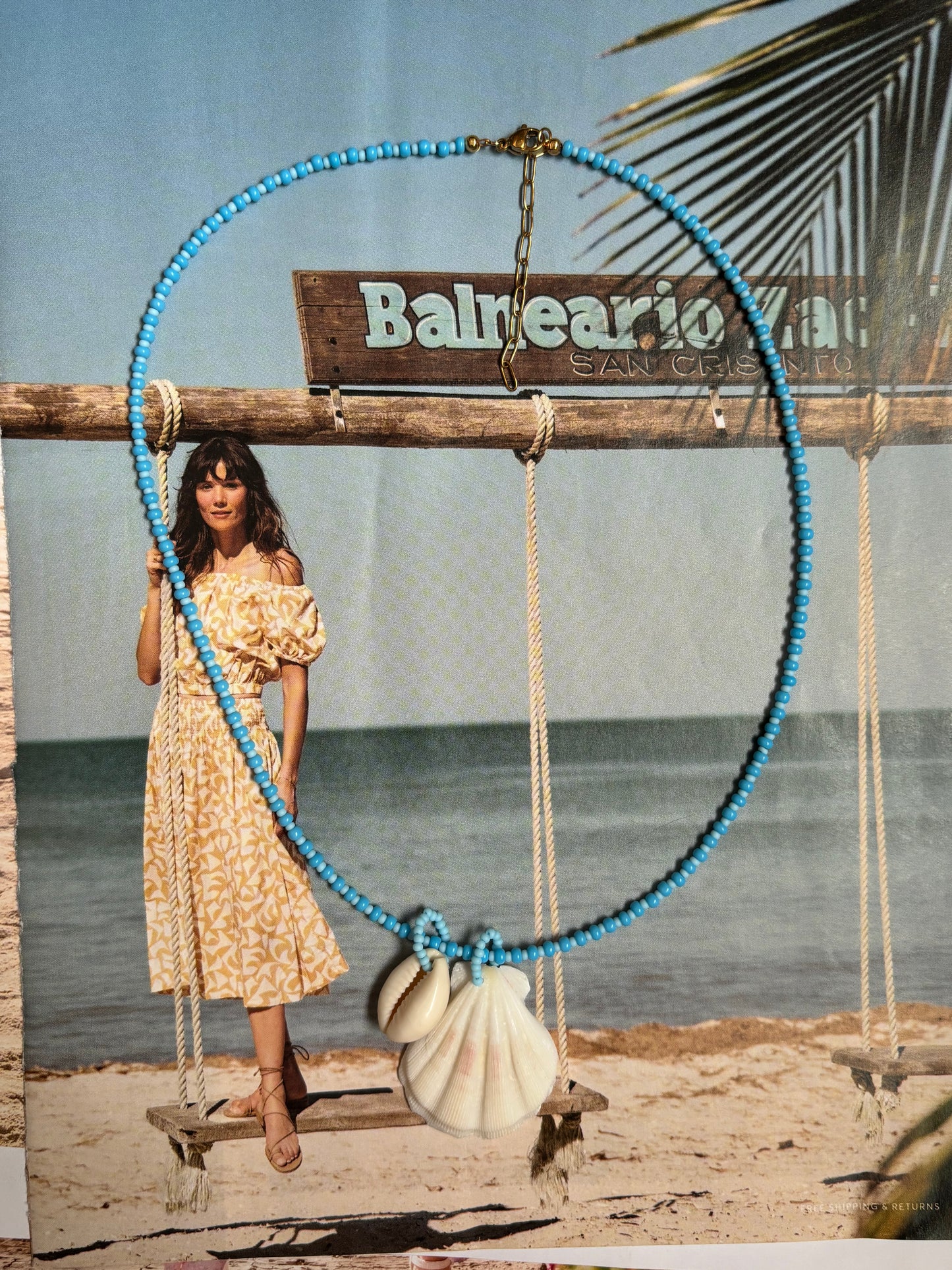 Coastal necklace with bright turquoise seed beads and three natural shell charms—scallop, cowrie, and conch—displayed in front of a beachy backdrop with ocean, swing set, and tropical style. Handmade by Soul Wave Beach Co with 18k gold-plated hardware, this fun and bold summer piece brings vacation vibes to everyday wear.
