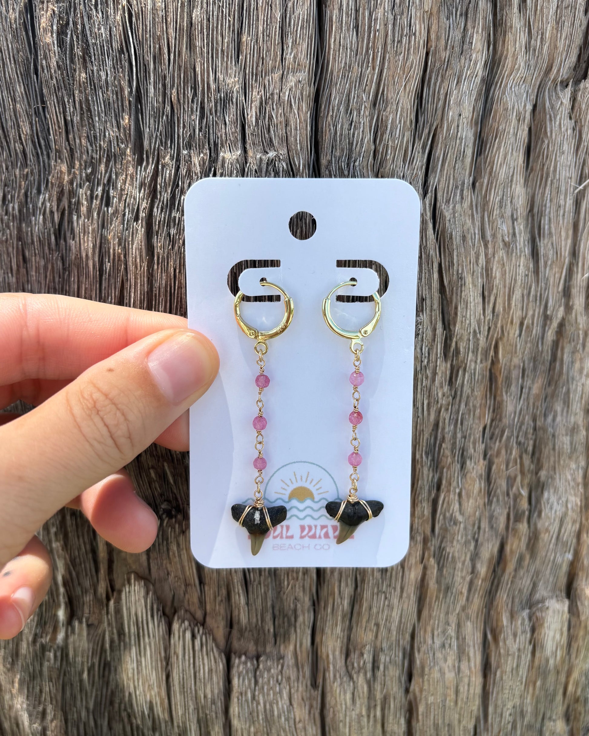 Shark tooth drop earrings with pink glass bead chain and fossilized shark teeth. Handcrafted with 14k gold-filled wire and suspended from gold-filled hoops. Earrings displayed on Soul Wave Beach Co. card held against driftwood. Beachy, handmade statement jewelry with a bold ocean vibe. Perfect for summer, surfers, and ocean lovers.