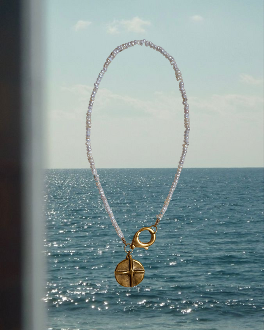 A delicate coastal necklace made with tiny freshwater pearls and finished with a gold coin pendant and oversized clasp, styled against a serene ocean backdrop. This beachy, nautical-inspired jewelry piece from Soul Wave Beach Co captures the essence of sun-soaked summers, seaside heritage, and minimalist ocean style. Perfect for layering or wearing solo, this handcrafted necklace reflects the Ocean Heritage collection’s vintage charm and timeless coastal elegance.