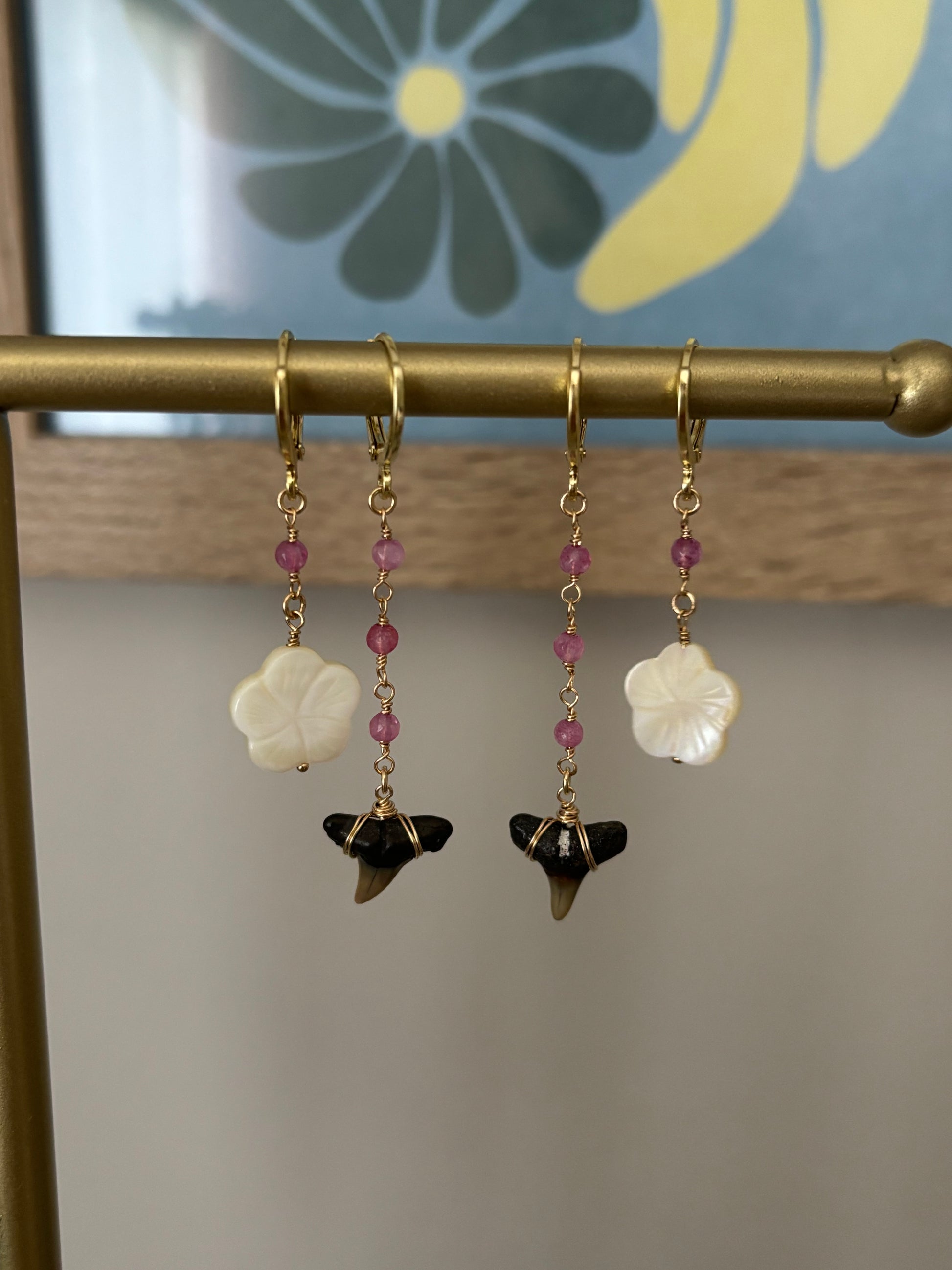 Tropical dangle earrings with white hibiscus flowers, black fossilized shark teeth, and pink gemstones hanging from 18k gold-plated hooks. Displayed on a brass bar with a floral wall art background. Handmade ocean-inspired earrings with gold and gemstone accents. Designed by Soul Wave Beach Co. for beach lovers and island getaways.