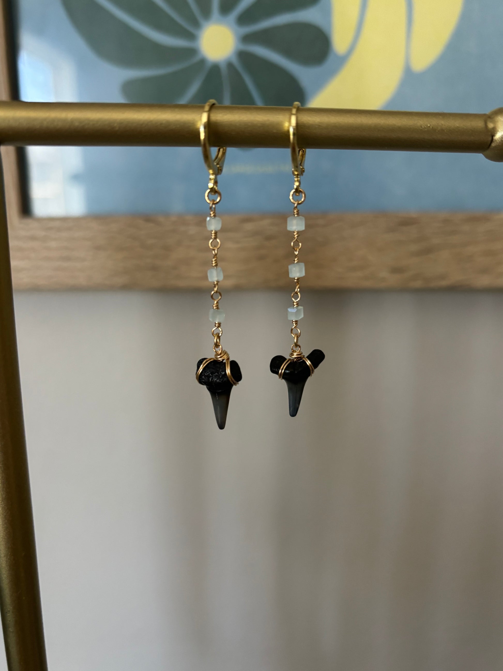 Shark tooth drop earrings with seafoam green square-cut glass bead chain, wrapped in 14k gold-filled wire and suspended from gold-filled hoops. Fossil shark teeth collected from South Florida. Lightweight and ocean-inspired, these handmade earrings by Soul Wave Beach Co. are perfect for beachwear, surfer style, and coastal gifting. Displayed on brass bar with tropical floral backdrop.
