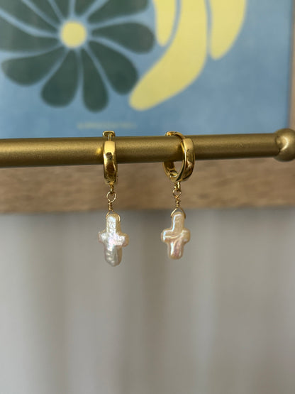 Pearl cross earrings hanging on a brass bar display. Earrings feature small cross-shaped freshwater pearls on 18k gold-plated huggie hoops, wire-wrapped with 14k gold-filled wire. Lightweight and beachy, ideal for summer outfits, bridal jewelry, and religious gift ideas. Coastal minimalist earrings by Soul Wave Beach Co. with tropical floral background.