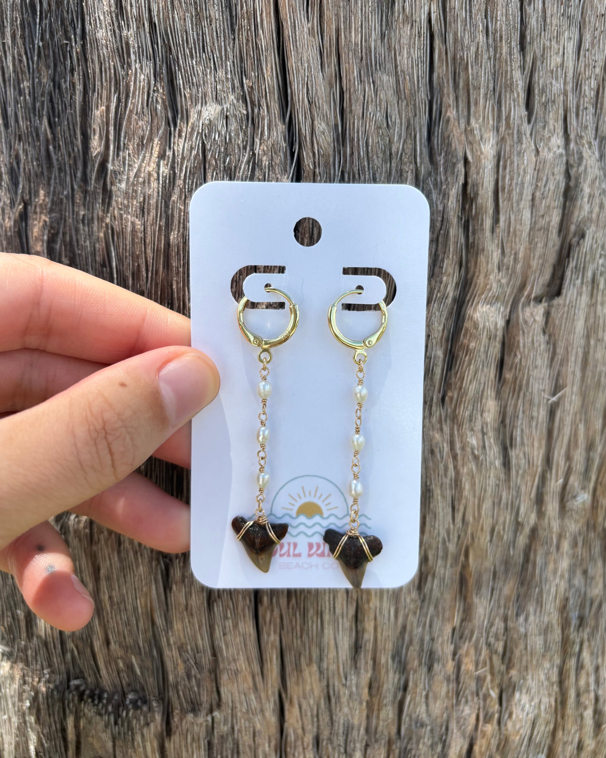 Handcrafted drop earrings featuring white pearl-like glass beads and real fossilized shark teeth. Wrapped in 14k gold-filled wire with matching gold-filled hoops. Displayed on branded Soul Wave Beach Co. earring card and photographed against a weathered driftwood background. Lightweight beach earrings with ocean-inspired style.