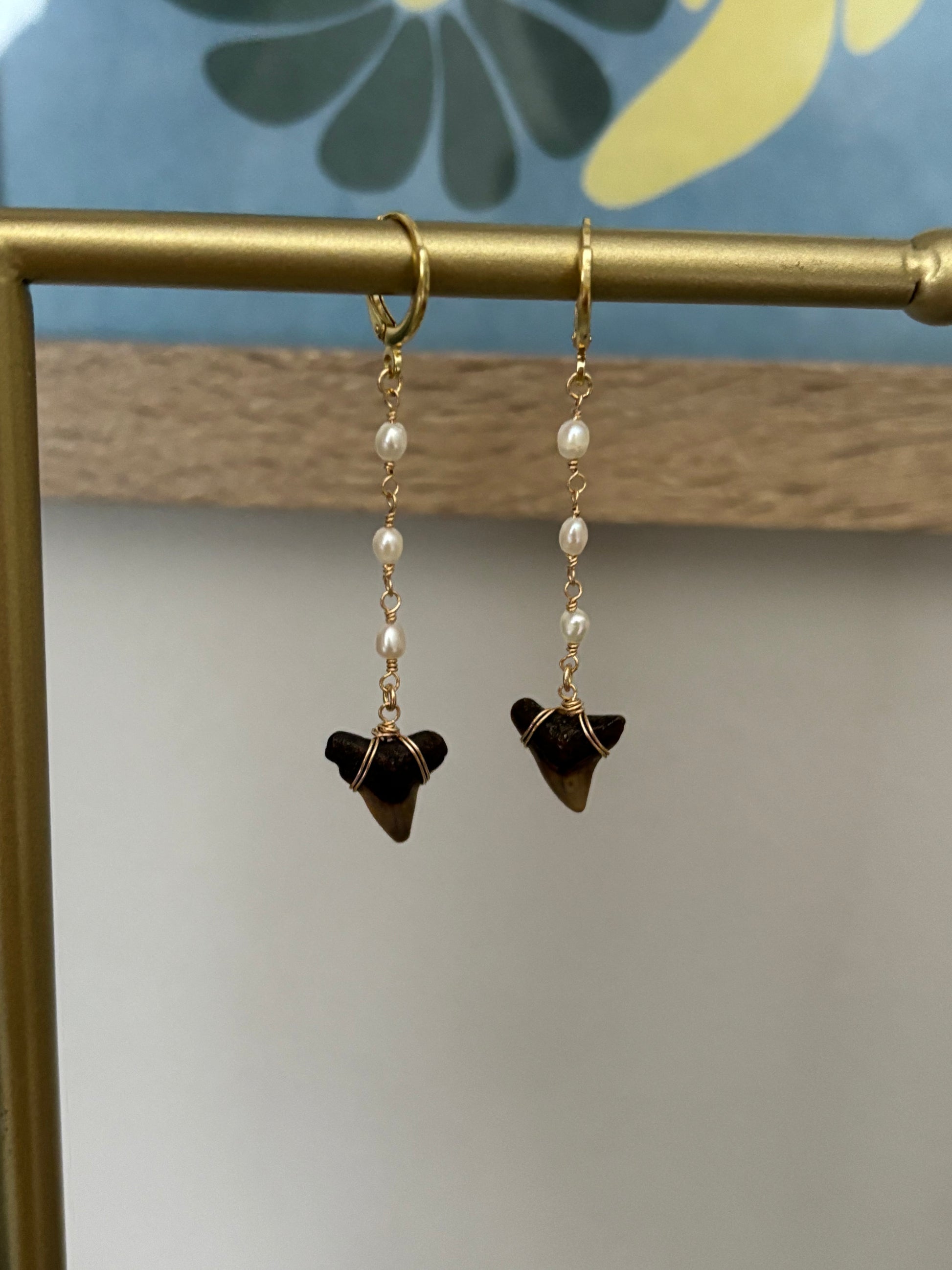 Shark tooth earrings with pearl-like white glass beaded chain, featuring hand-collected fossil shark teeth wire-wrapped in 14k gold-filled wire. Suspended from gold-filled hoops for a polished beachy finish. Handmade by Soul Wave Beach Co., these earrings are perfect for ocean lovers, beach brides, or coastal everyday wear. Displayed on a brass jewelry bar with floral background.