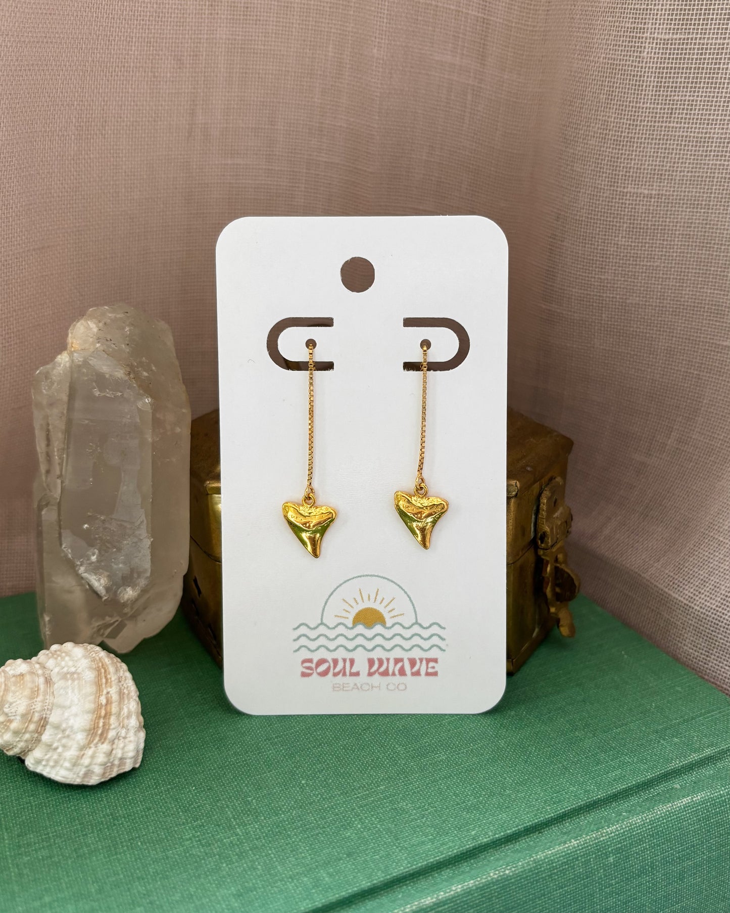 Pair of dainty gold shark tooth threader earrings on a branded Soul Wave Beach Co. card. Earrings are made from 14k gold filled materials with mini shark tooth charms hanging from delicate threader chains. Styled with seashell, crystal, and vintage box on a green fabric surface with a blush-toned backdrop. Perfect coastal jewelry for beach lovers, ocean-inspired gifts, surfer girls, or summer accessories. Lightweight, minimalist, and handmade.