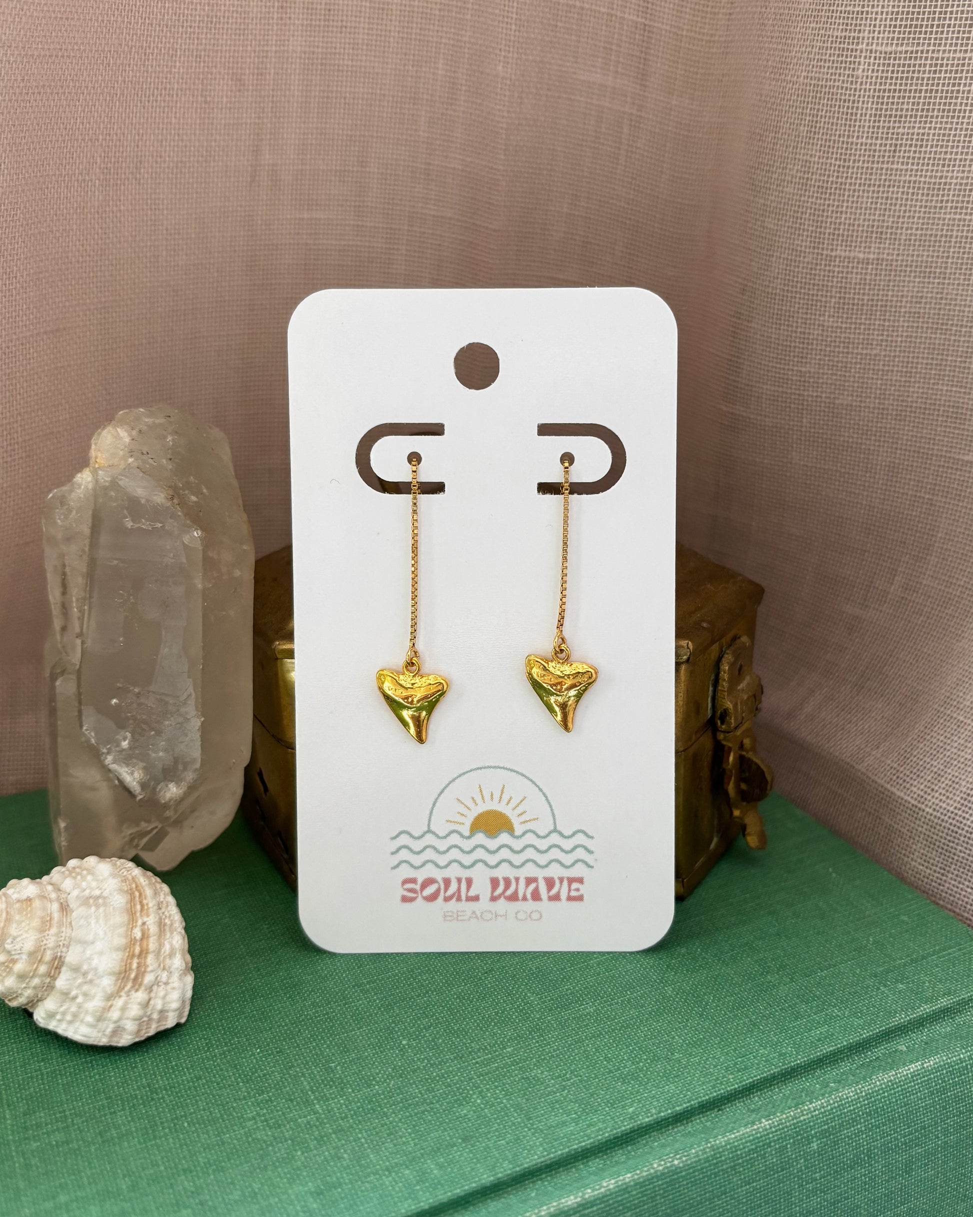 Pair of dainty gold shark tooth threader earrings on a branded Soul Wave Beach Co. card. Earrings are made from 14k gold filled materials with mini shark tooth charms hanging from delicate threader chains. Styled with seashell, crystal, and vintage box on a green fabric surface with a blush-toned backdrop. Perfect coastal jewelry for beach lovers, ocean-inspired gifts, surfer girls, or summer accessories. Lightweight, minimalist, and handmade.