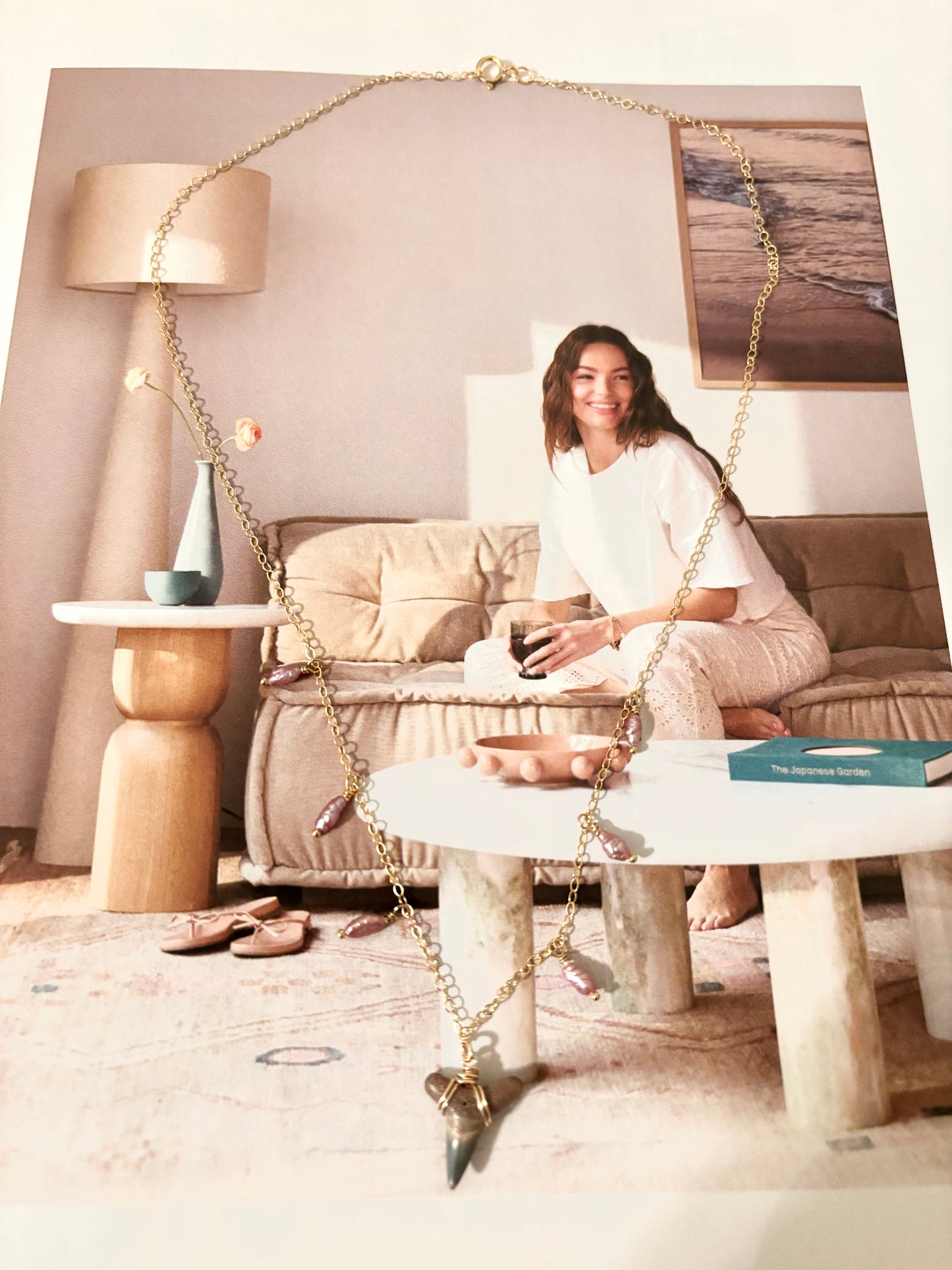 Flat lay of a handcrafted shark tooth necklace made from 14k gold filled sterling silver chain with antique purple freshwater rice pearls. Necklace is styled on a lifestyle magazine showing a relaxed woman in cozy loungewear. Coastal-inspired, beachy boho jewelry handmade by Soul Wave Beach Co. Perfect gift for ocean lovers, summer vibes, or sea-themed accessories. Great for everyday coastal chic style.