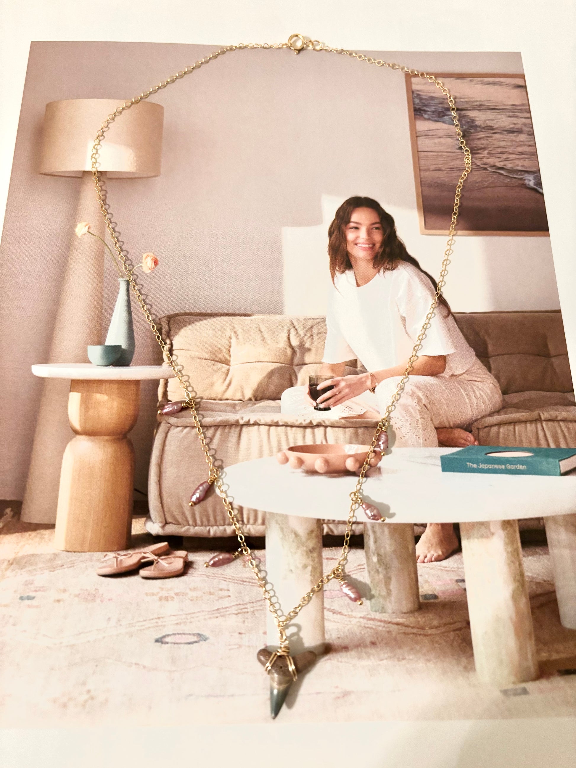 Flat lay of a handcrafted shark tooth necklace made from 14k gold filled sterling silver chain with antique purple freshwater rice pearls. Necklace is styled on a lifestyle magazine showing a relaxed woman in cozy loungewear. Coastal-inspired, beachy boho jewelry handmade by Soul Wave Beach Co. Perfect gift for ocean lovers, summer vibes, or sea-themed accessories. Great for everyday coastal chic style.