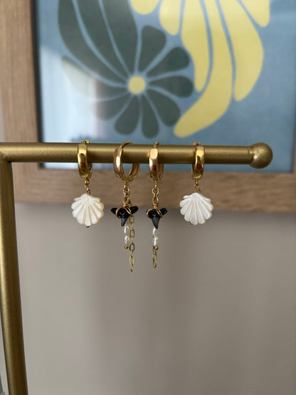 Mother of pearl shell-shaped earrings displayed on a gold brass bar with floral wall art in the background. Features gold-plated hoops and 14k gold-filled wire wrapped around carved shell charms. Coastal minimal jewelry handmade by Soul Wave Beach Co. Perfect for summer styling, beachy weddings, ocean lovers, and boho chic accessorizing in white and gold.