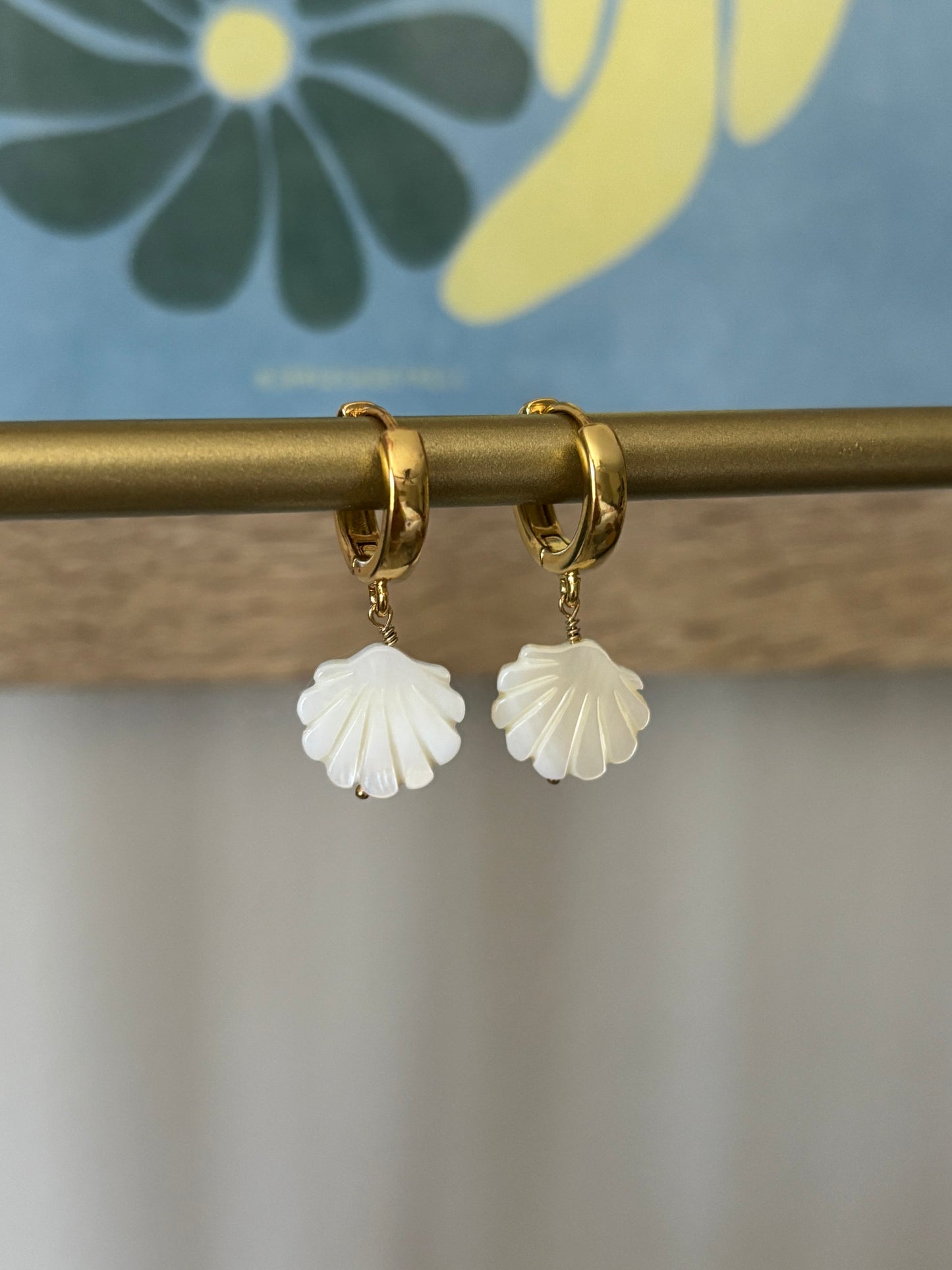 Close-up of mother of pearl shell earrings hanging on 18k gold-plated huggie hoops with 14k gold-filled wire. Displayed on brass jewelry bar with colorful floral background. Lightweight carved shell charms in a fan shape. Handmade coastal-inspired earrings perfect for summer, beach brides, or boho outfits. Soul Wave Beach Co. ocean-themed jewelry in natural white and gold tones.