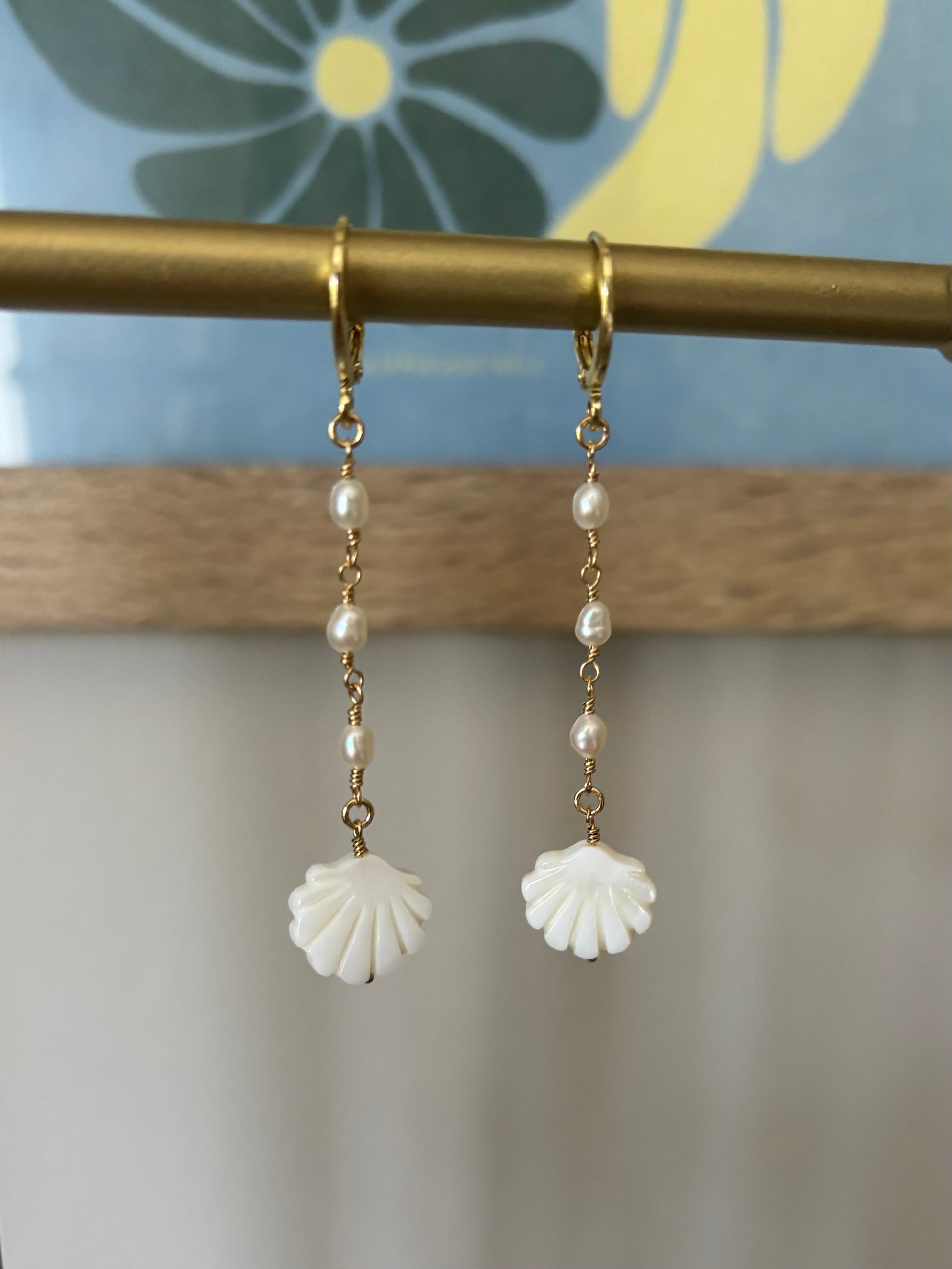 Mother of pearl shell drop earrings hanging from a brass display bar. Each earring features a luminous freshwater pearl and hand-carved shell charm wire-wrapped in 14k gold-filled metal. Lightweight ocean-inspired jewelry handmade by Soul Wave Beach Co. Ideal for coastal outfits, beach vacations, and boho summer styling.