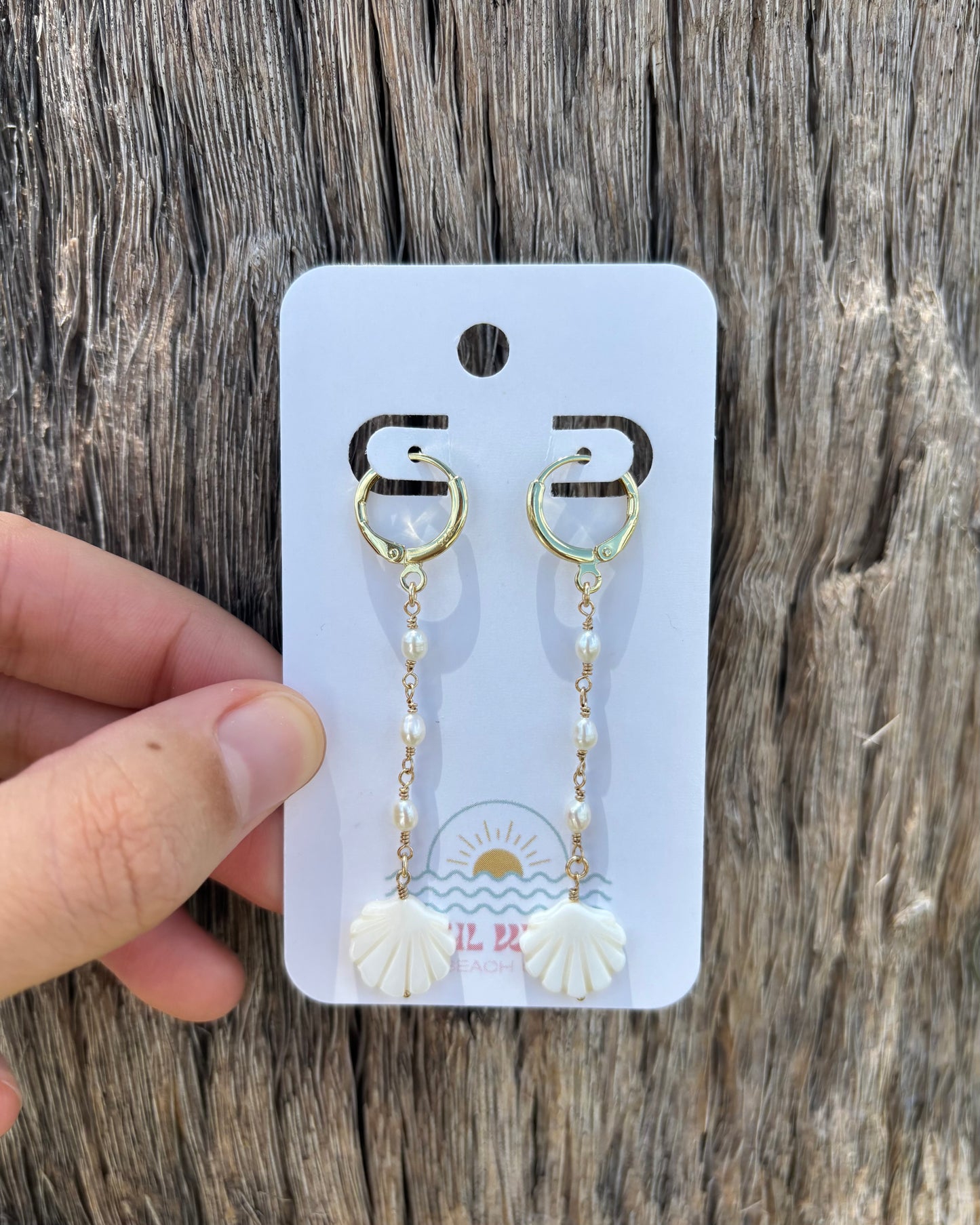 Hand holding mother of pearl shell drop earrings on Soul Wave Beach Co. earring card. Features carved shell charms and freshwater pearls wrapped in 14k gold-filled wire. Lightweight and beachy dangle earrings handmade for summer, bridal, or everyday resort wear. Photographed against rustic driftwood background with natural lighting for a coastal aesthetic.