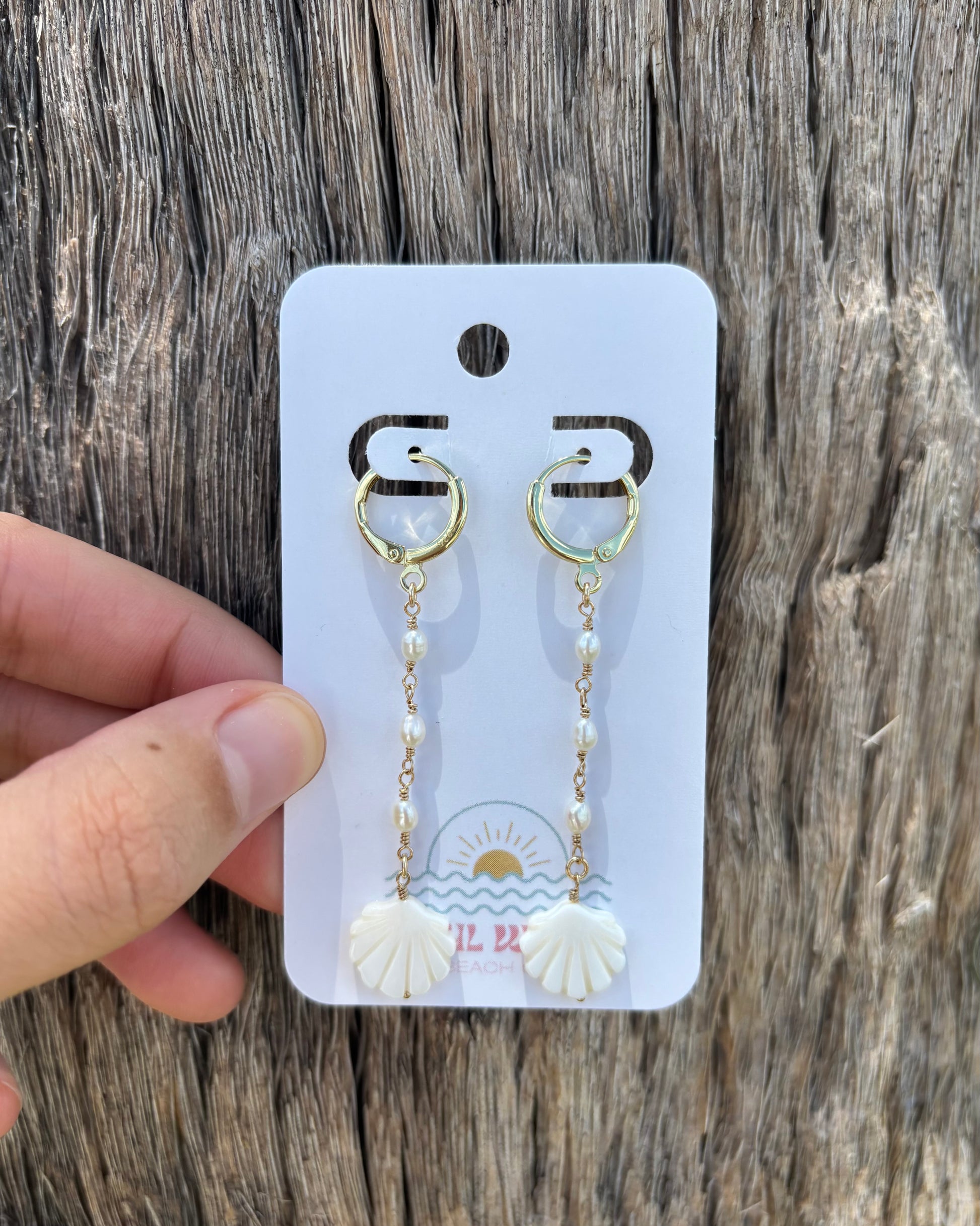 Hand holding mother of pearl shell drop earrings on Soul Wave Beach Co. earring card. Features carved shell charms and freshwater pearls wrapped in 14k gold-filled wire. Lightweight and beachy dangle earrings handmade for summer, bridal, or everyday resort wear. Photographed against rustic driftwood background with natural lighting for a coastal aesthetic.