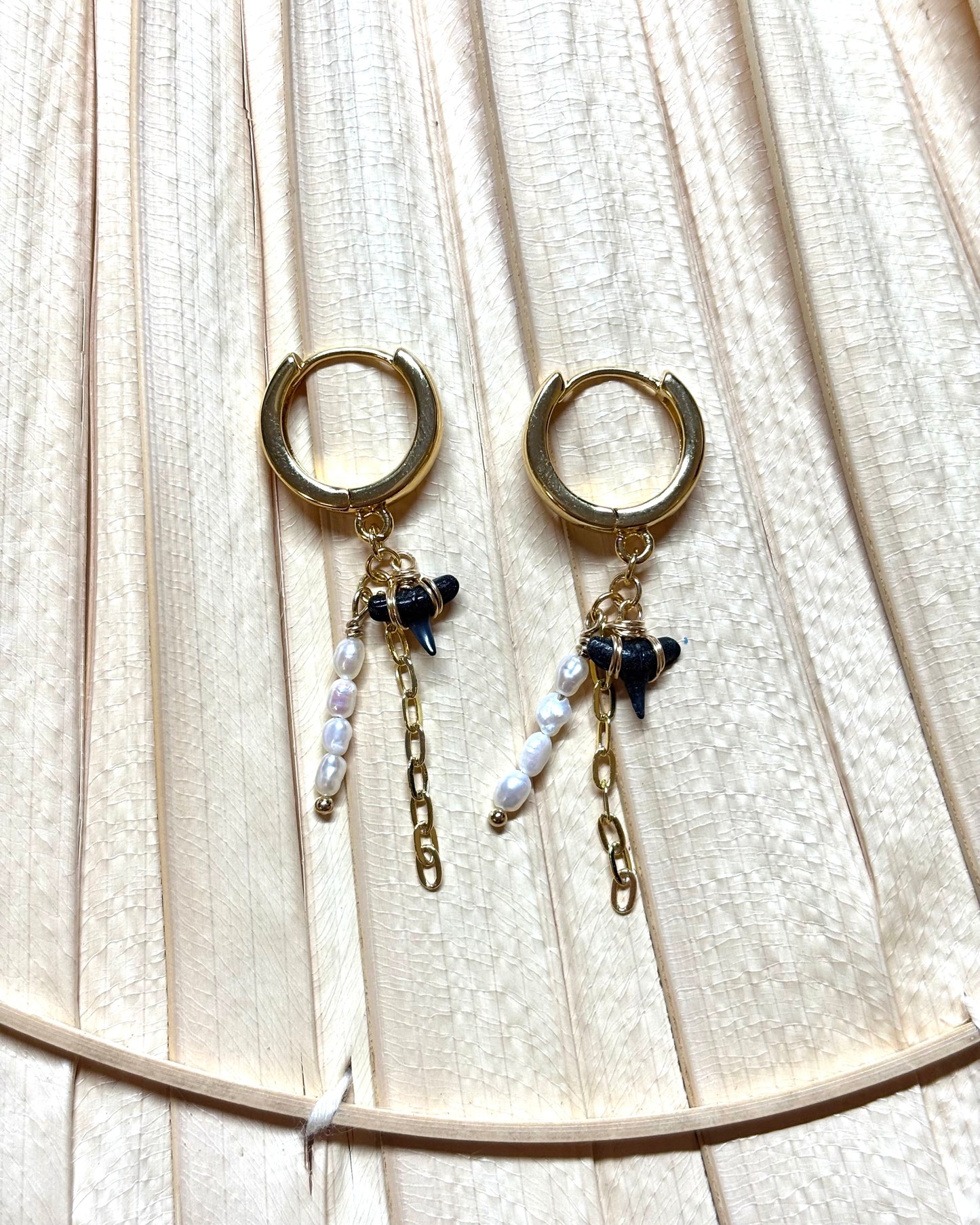 Gold hoop earrings with real hand-collected shark teeth, freshwater pearls, and paperclip chain dangles displayed on a neutral palm leaf background. These coastal-inspired dangle earrings blend ocean charm with bold boho style, making them perfect for beach lovers and statement jewelry fans. Handmade and best-selling, ideal for tropical vacations, summer outfits, or everyday surf-inspired fashion.