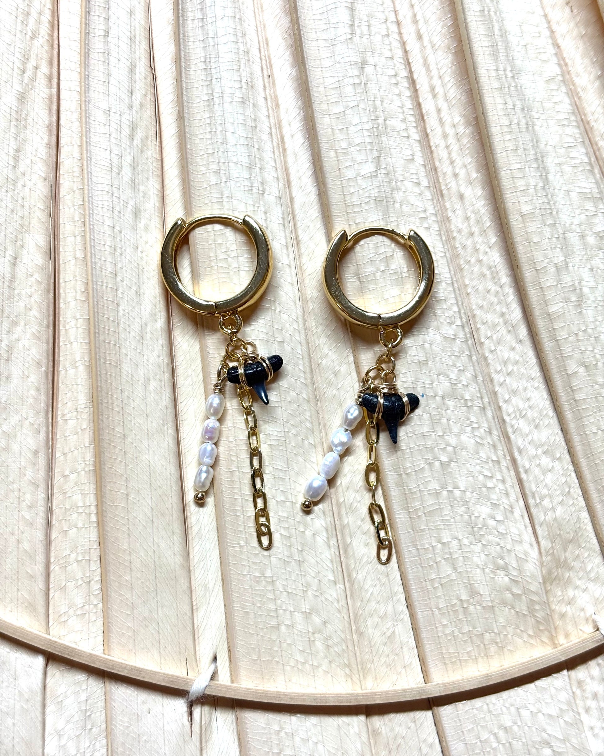 Gold hoop earrings with real hand-collected shark teeth, freshwater pearls, and paperclip chain dangles displayed on a neutral palm leaf background. These coastal-inspired dangle earrings blend ocean charm with bold boho style, making them perfect for beach lovers and statement jewelry fans. Handmade and best-selling, ideal for tropical vacations, summer outfits, or everyday surf-inspired fashion.