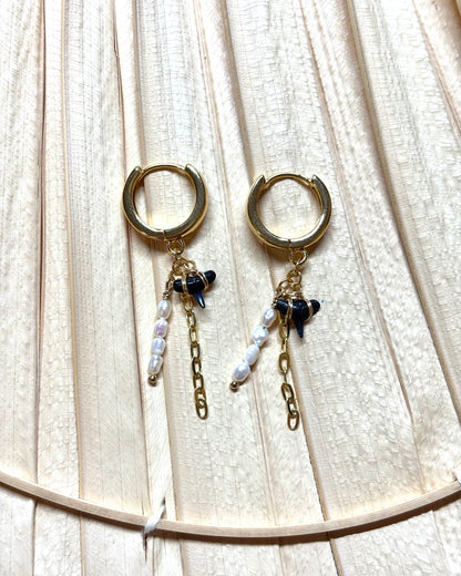 Gold hoop earrings with real hand-collected shark teeth, freshwater pearls, and paperclip chain dangles displayed on a neutral palm leaf background. These coastal-inspired dangle earrings blend ocean charm with bold boho style, making them perfect for beach lovers and statement jewelry fans. Handmade and best-selling, ideal for tropical vacations, summer outfits, or everyday surf-inspired fashion.