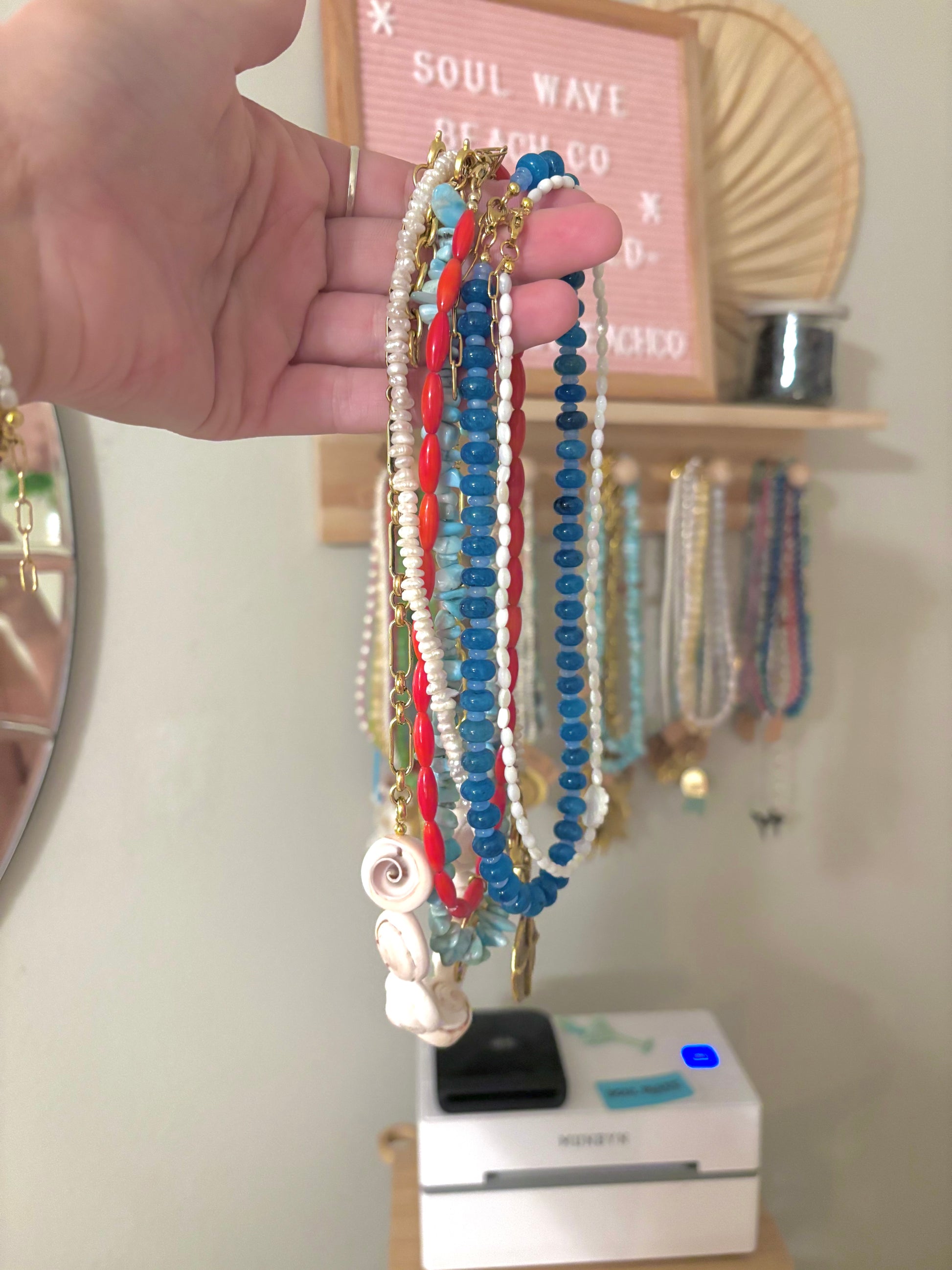 Hand holding a bundle of coastal-inspired beaded necklaces featuring aquamarine, coral red, freshwater pearls, white seed beads, and shell charms. Each necklace is handmade with gold-plated findings, perfect for a beachy, boho summer look. Soul Wave Beach Co branding sign in background.