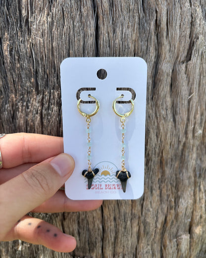 Seafoam green glass beaded shark tooth earrings with fossil teeth wrapped in 14k gold-filled wire. Hanging from gold-filled hoops on Soul Wave Beach Co. earring card. Natural ocean tones and beachy drop design, perfect for summer styling, handmade jewelry gifting, or everyday coastal wear. Set against driftwood for a seaside aesthetic.