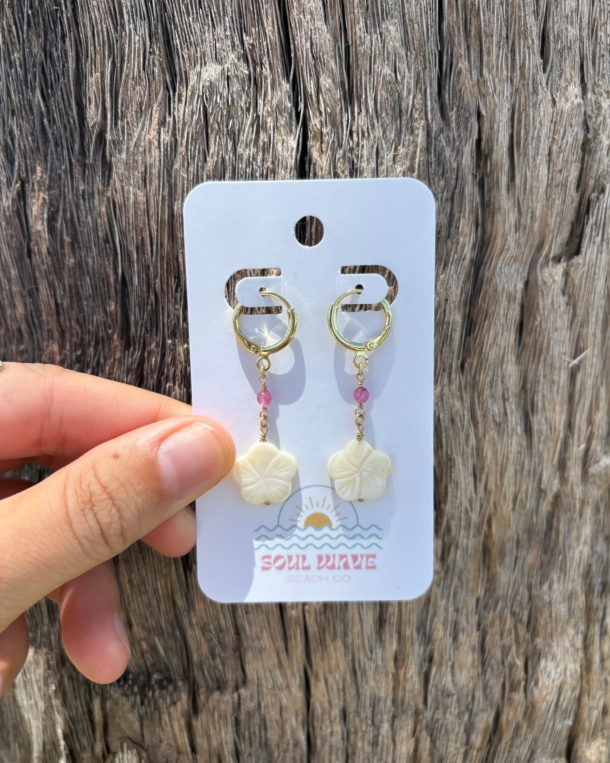 Hand holding Hibiscus Dangle Earrings on Soul Wave Beach Co. card. Features carved mother of pearl hibiscus flowers, black shark teeth, and pink gemstone beads on gold-filled wire and 18k gold-plated hooks. Handmade coastal statement earrings, perfect for tropical vacations, beach weddings, or summer styling. Displayed against rustic driftwood.