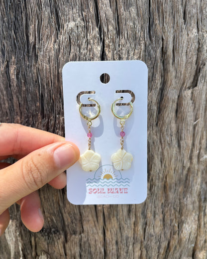 Hand holding Hibiscus Dangle Earrings on Soul Wave Beach Co. card. Features carved mother of pearl hibiscus flowers, black shark teeth, and pink gemstone beads on gold-filled wire and 18k gold-plated hooks. Handmade coastal statement earrings, perfect for tropical vacations, beach weddings, or summer styling. Displayed against rustic driftwood.