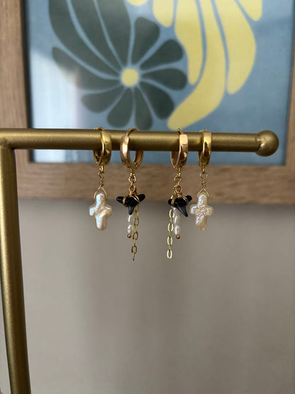Two pairs of mismatched gold huggie hoop earrings displayed on a brass jewelry bar. Features two pearl cross earrings and two black shark tooth earrings with freshwater pearls and gold chain dangles. All earrings are handcrafted with 14k gold-filled wire and 18k gold-plated hoops. Designed by Soul Wave Beach Co., this coastal jewelry blends beachy elegance with bold ocean vibes. Set against a floral art background for a modern tropical aesthetic.