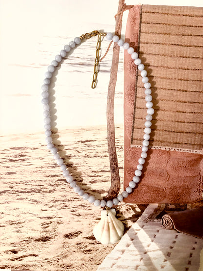 Light blue beaded necklace with natural scallop shell pendant, photographed against a beachy backdrop with sand and driftwood. Handmade by Soul Wave Beach Co, this ocean-inspired piece features blue aquamarine beads, a 18k gold-filled clasp, and adjustable extender chain. A soft coastal necklace perfect for layering or making a relaxed summer statement.