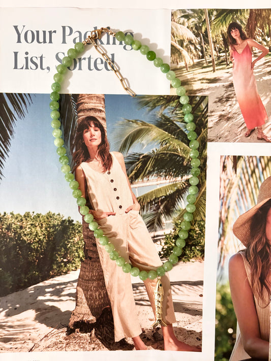Tropical green gemstone necklace made with polished aventurine beads and a gold-toned fish charm pendant, displayed over a beachy fashion magazine backdrop with palm trees and sand. Features a 18k gold-plated clasp and extender chain. Handmade by Soul Wave Beach Co, this beach-inspired necklace blends playful charm with ocean-toned elegance—perfect for summer or resort styling.
