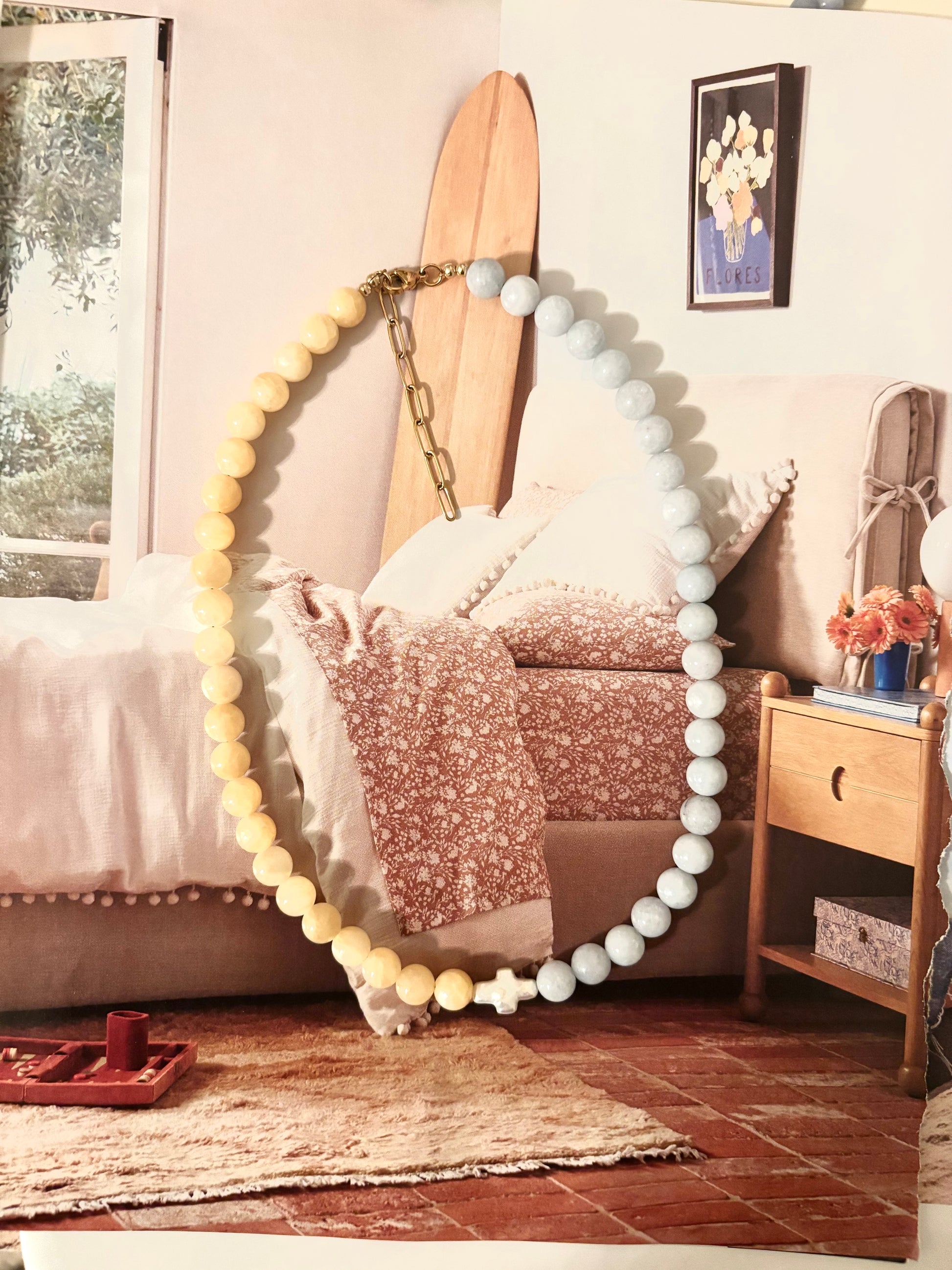 Pastel beaded necklace with yellow jade and light blue aquamarine beads, finished with a pearl cross charm at center. Displayed on a bed with coastal decor and a mini surfboard, this necklace is handmade by Soul Wave Beach Co. Features 18k gold-plated clasp and extender. A peaceful, soft-toned piece perfect for beach days, Sunday mornings, or layered coastal looks.