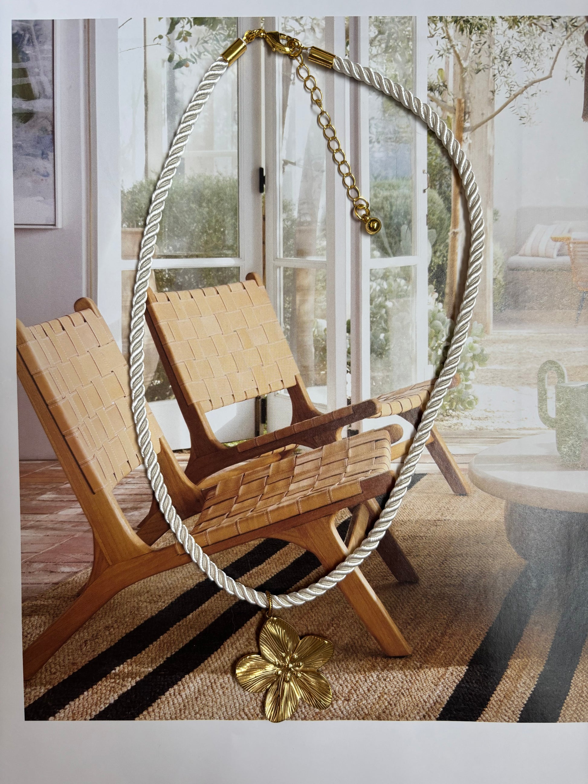 Chunky twisted white cord necklace with a large gold hibiscus-style flower pendant, styled on a neutral modern beach interior with woven chairs and natural light. Handmade by Soul Wave Beach Co, this tropical statement necklace features gold-plated hardware and an adjustable extender chain. A bold, retro-inspired piece for vacation outfits, beach weddings, or summer resort looks.
