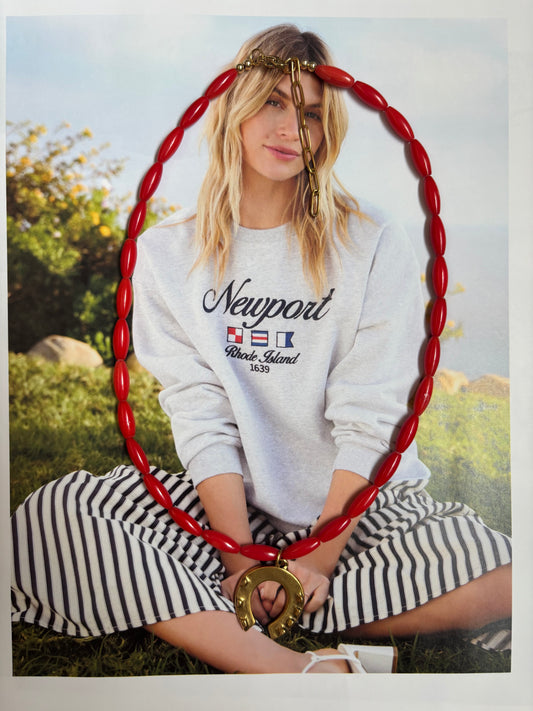 High Tide Luck Necklace with red coral beads and a gold horseshoe charm, styled over a coastal-inspired outfit with a Newport sweatshirt and striped skirt. Handmade by Soul Wave Beach Co, this statement necklace features natural coral, a 18k gold-plated clasp, and adjustable extender. A lucky charm with bold red tones—perfect for nautical, beachy, or resort styling.