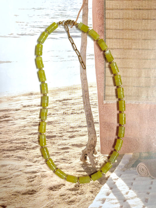 Matcha green beaded necklace with chunky glass cylinder beads and gold-filled accents, styled against a beach background with sand, driftwood, and towel. Handmade by Soul Wave Beach Co, this bold summer necklace features an adjustable 14k gold-filled clasp and extender chain. A colorful statement piece with coastal charm, perfect for layering or wearing solo.